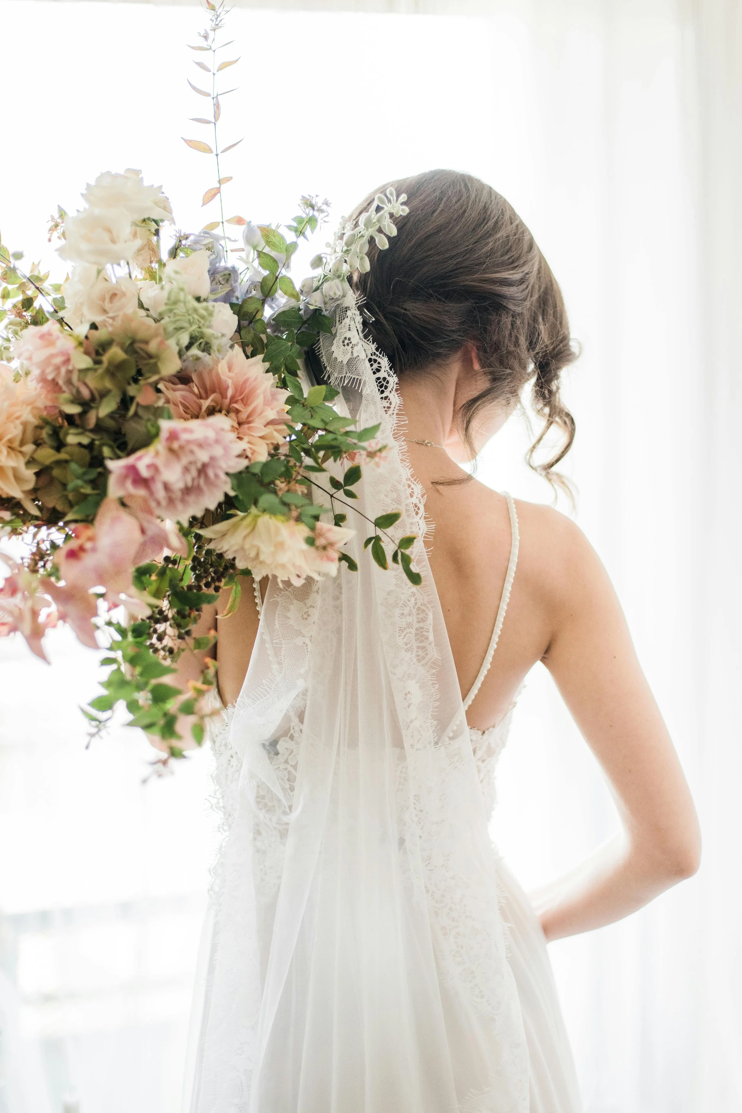 A bride with brown hair in a lace wedding dress and veil, holding a large bouquet of pink, white, and green flowers near a window with sheer curtains.