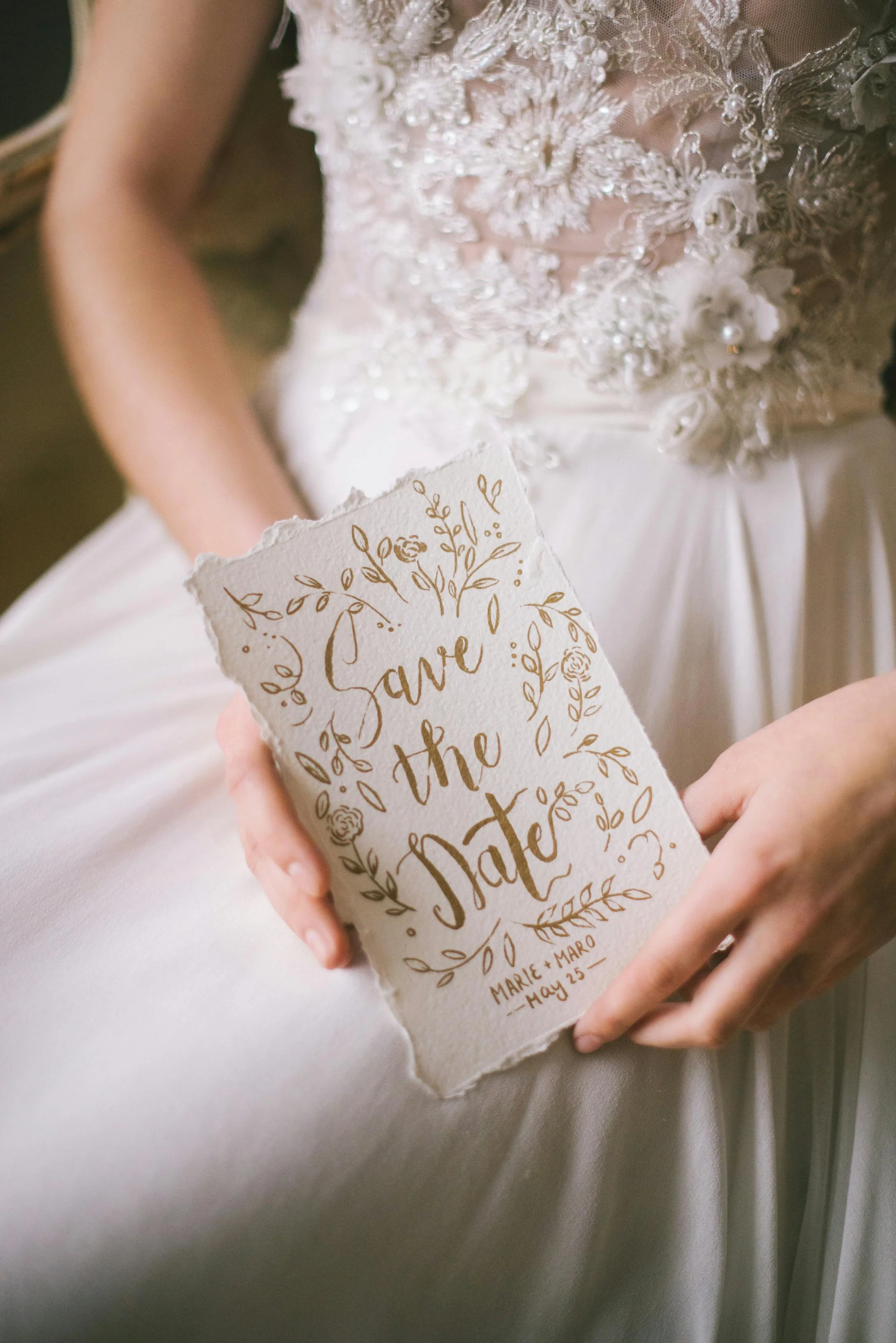 A bride in a wedding dress holding a handmade 'Save the Date' card with floral designs and the names Marie and Maro, dated May 25.
