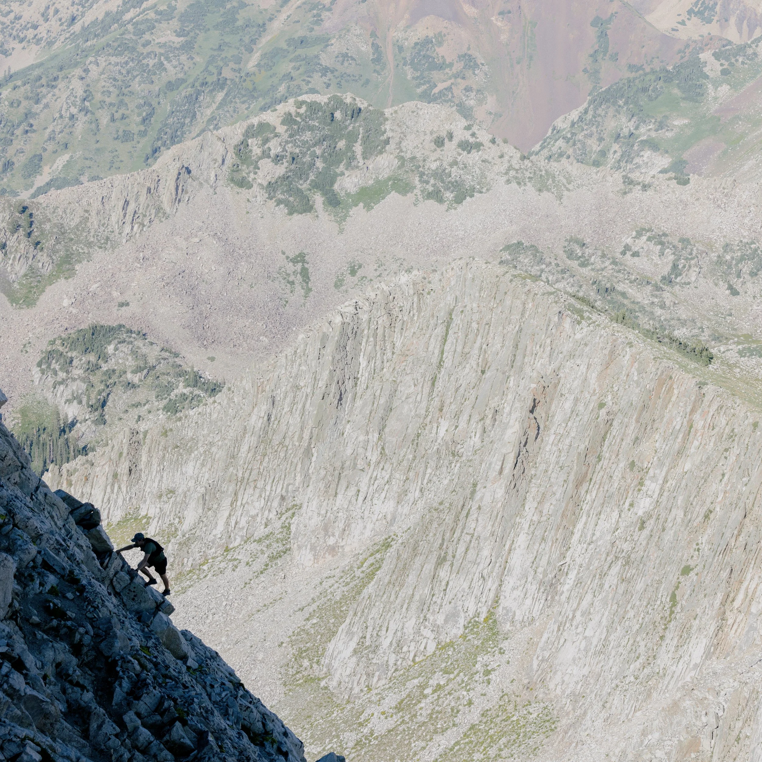 A person climbing a steep rocky mountain slope near a rugged, cratered mountain landscape with green patches in the background.