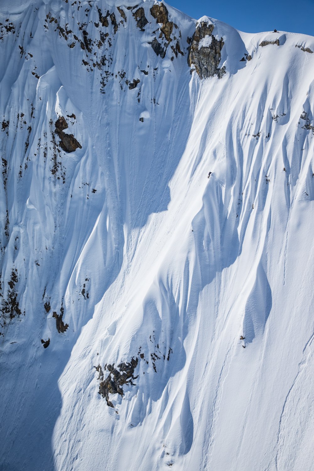 A steep snow-covered mountain slope with visible rock outcrops and snow avalanches falling down the face against a clear blue sky.