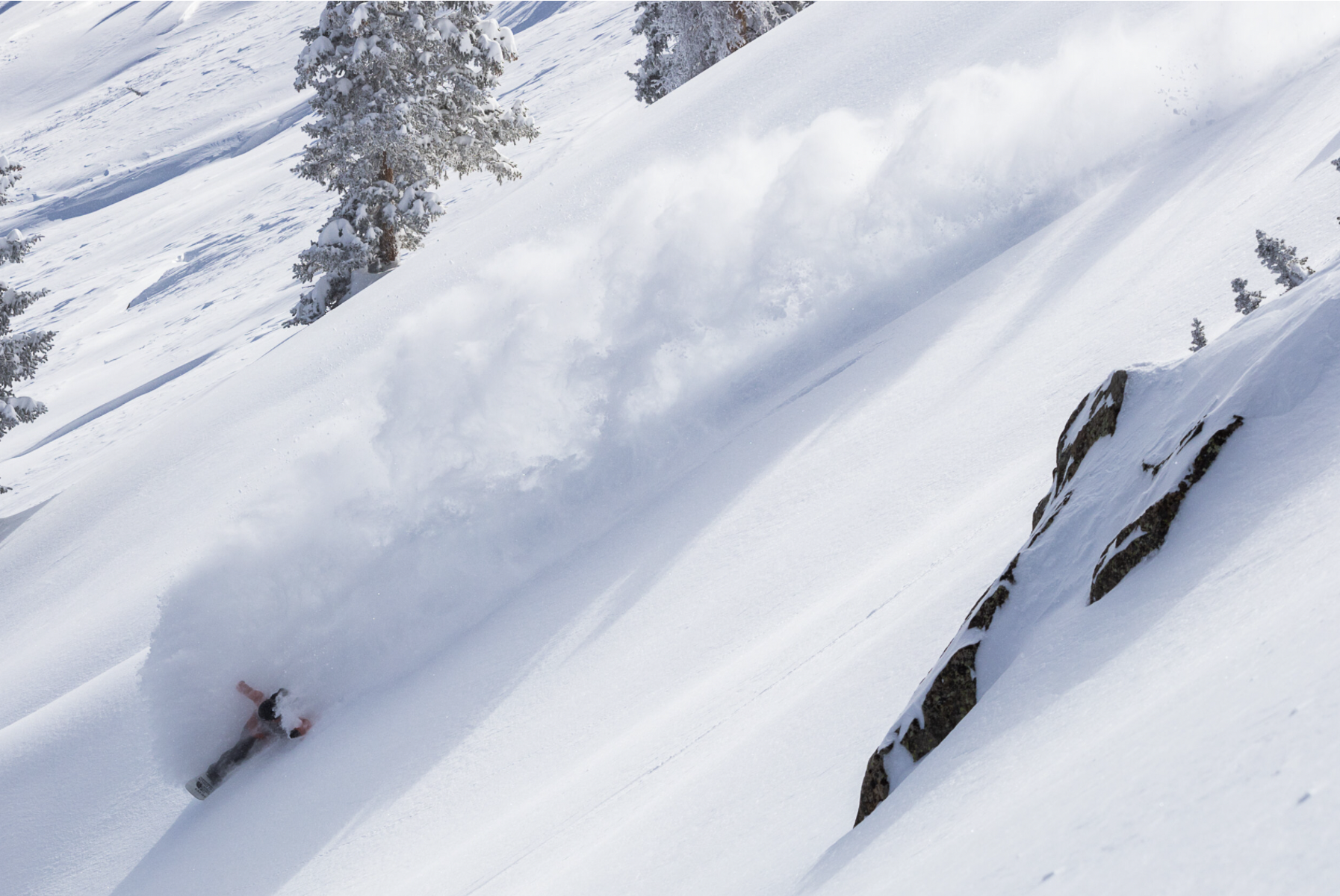 A person skiing down a snowy mountain slope surrounded by snow-covered trees and rocks, with snow spray created by the turn.