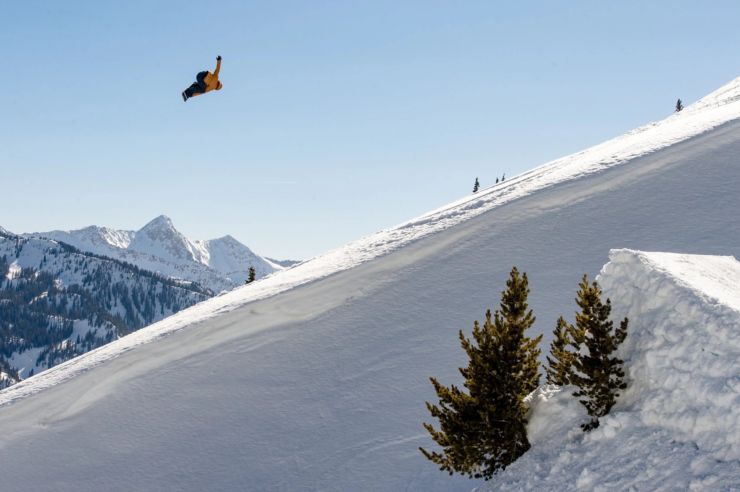 A person in an orange jacket is mid-air performing a backflip on a snowy mountain slope, with snow-covered peaks and a clear blue sky in the background.