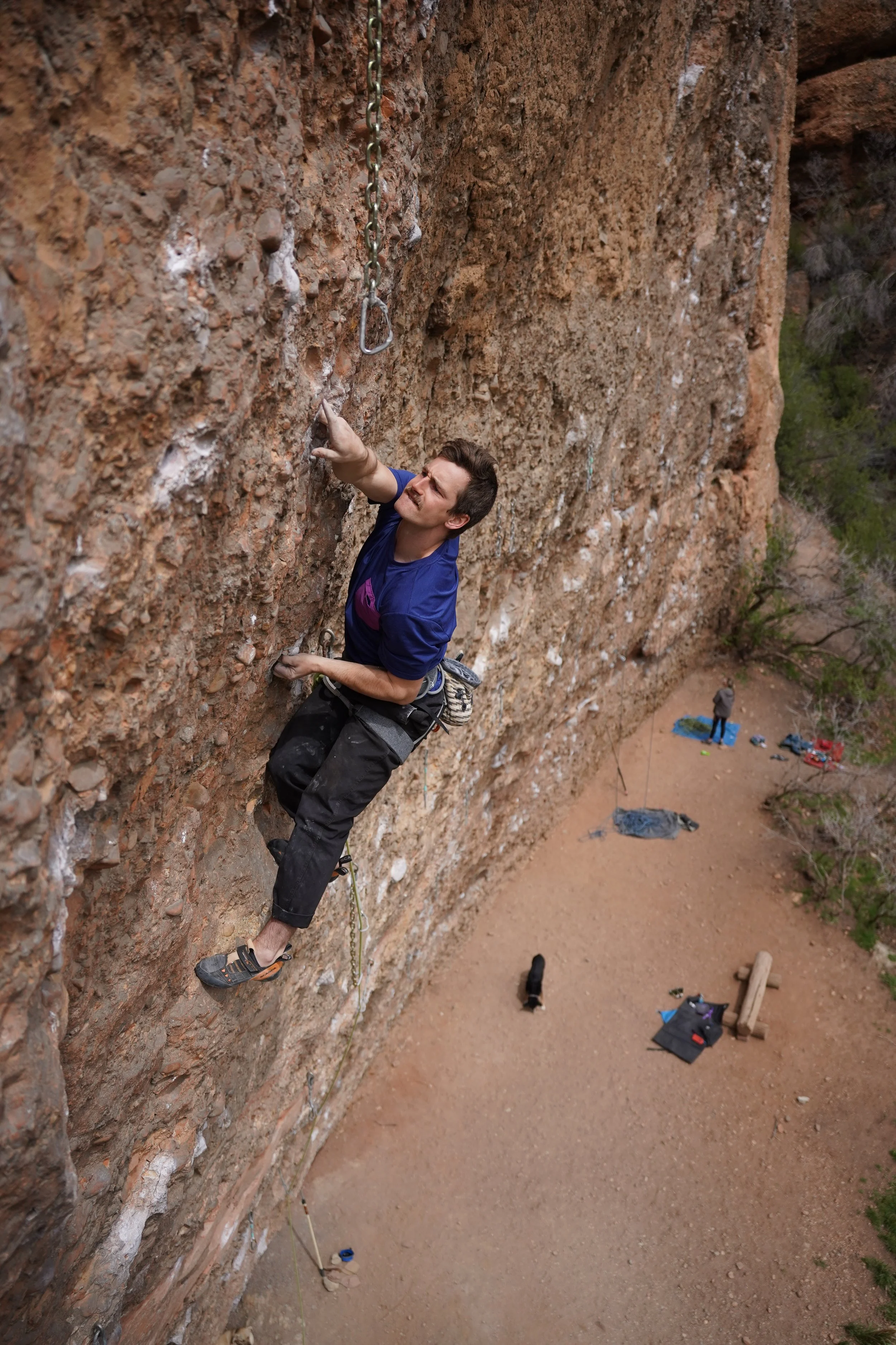 A man rock climbing on a tall, vertical, reddish-brown cliff face, with a climbing harness and notes on the ground below.
