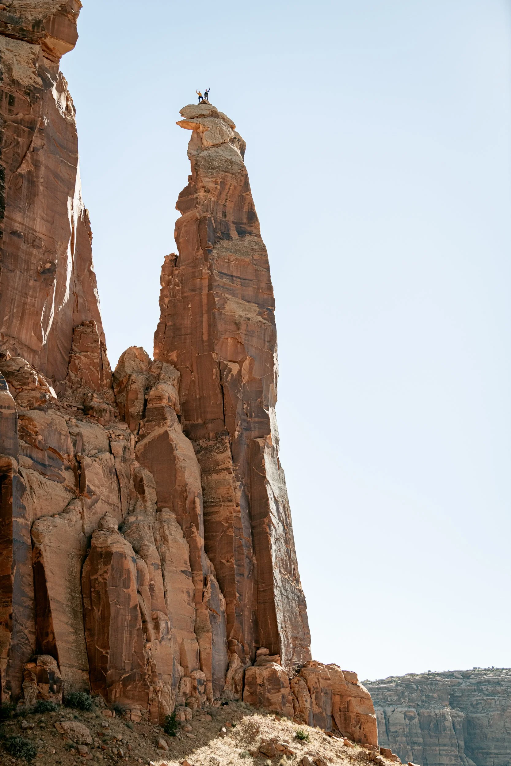 Two people standing on top of a tall rock formation in a desert landscape, with a clear blue sky overhead.
