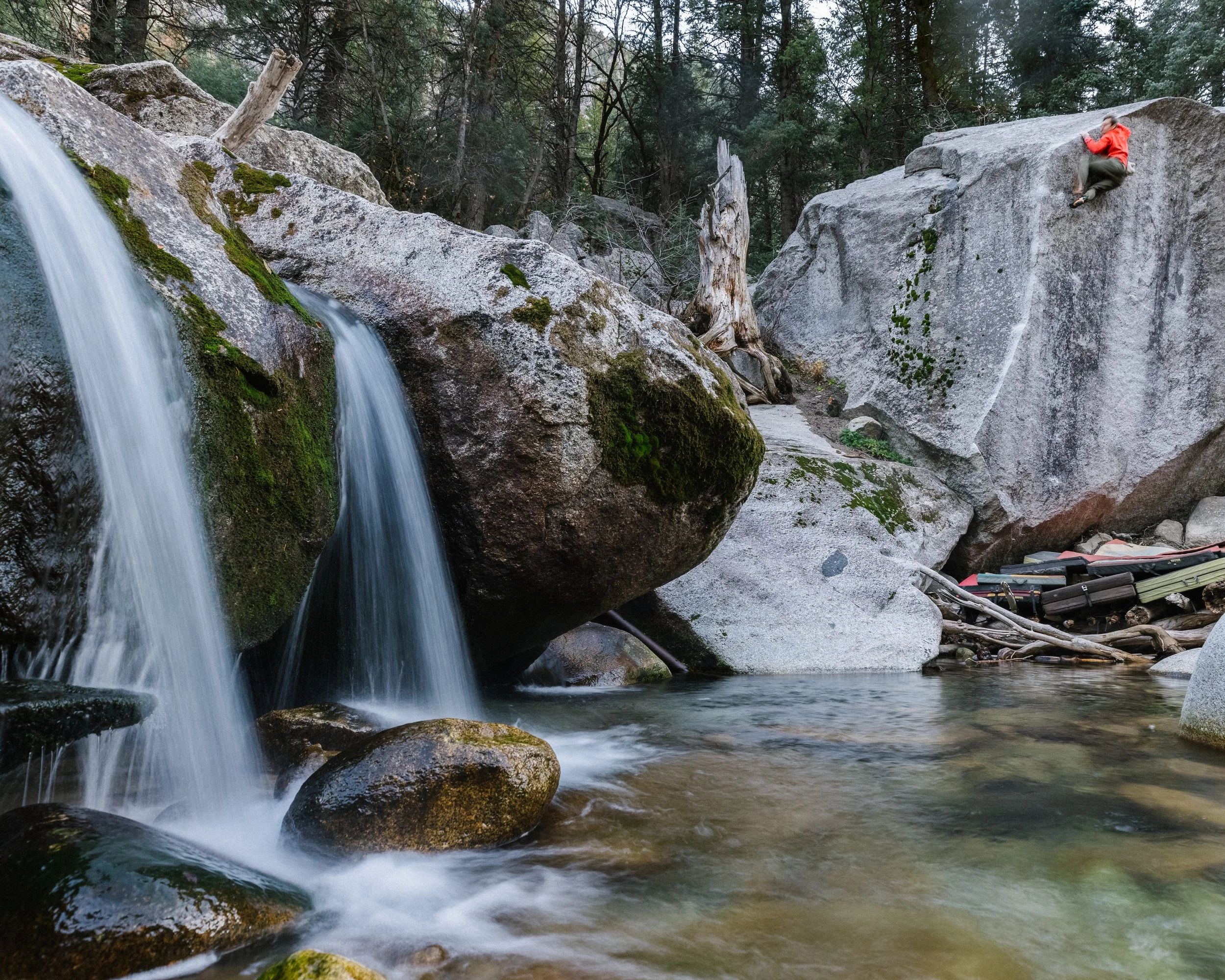 A person wearing a red jacket climbs a large granite rock near a waterfall in a forested area.
