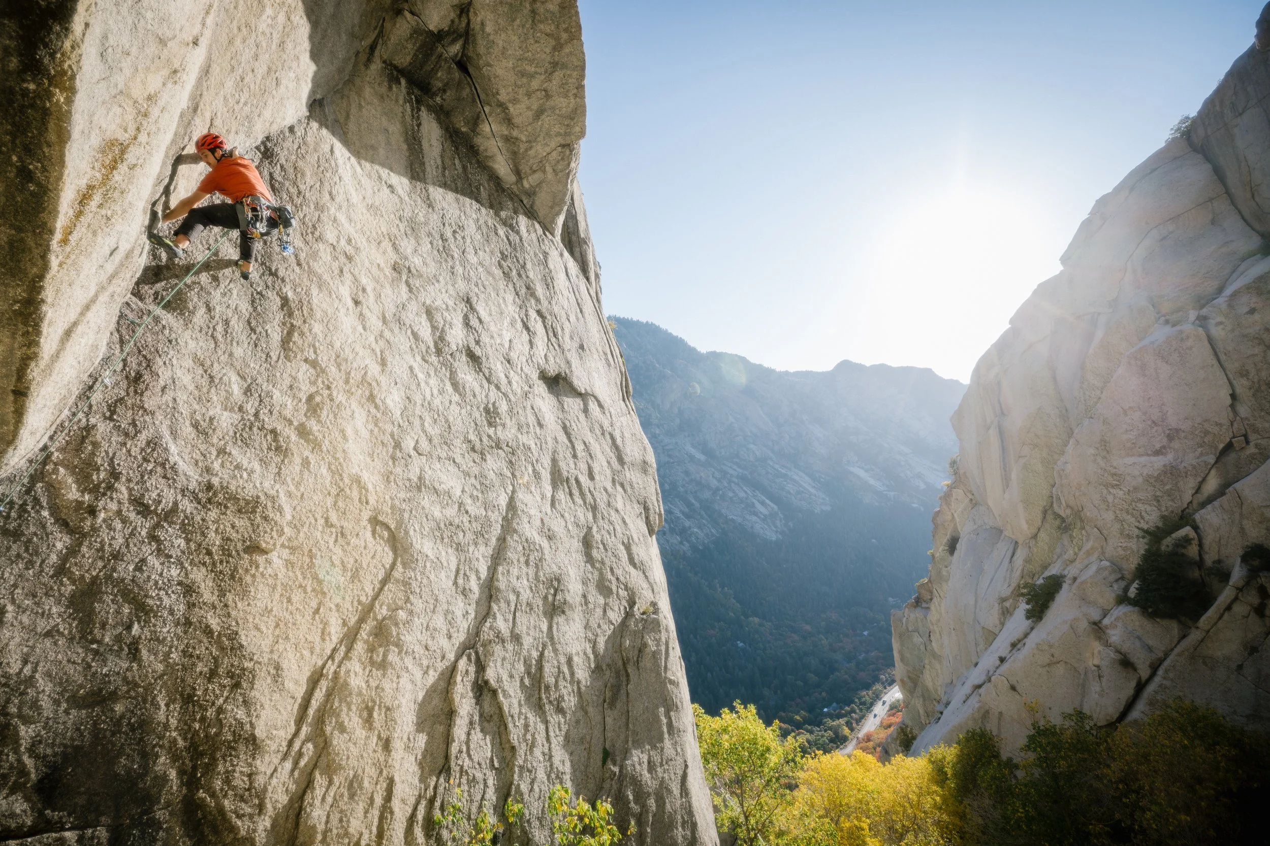 A person in an orange shirt and harness climbing a tall granite rock face amid a mountain landscape with sunlight and clear sky.
