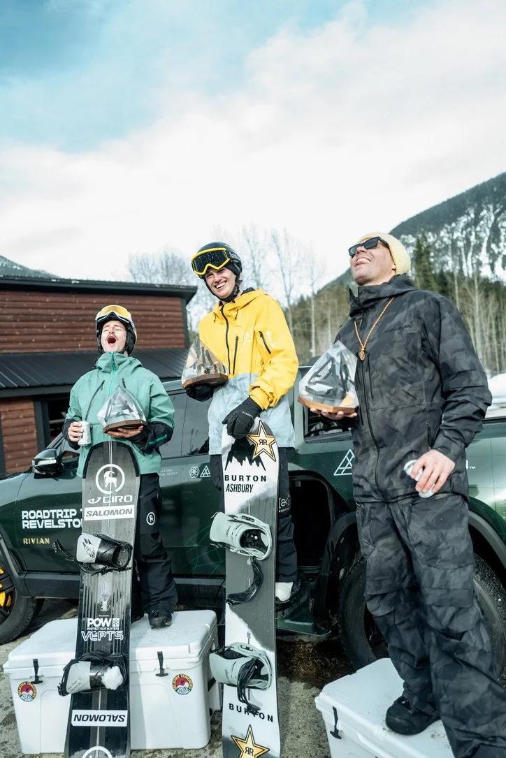 Three people in winter gear standing on a podium outdoors after a snowboarding competition, holding trophies and with snowboards, with a car and mountains in the background.