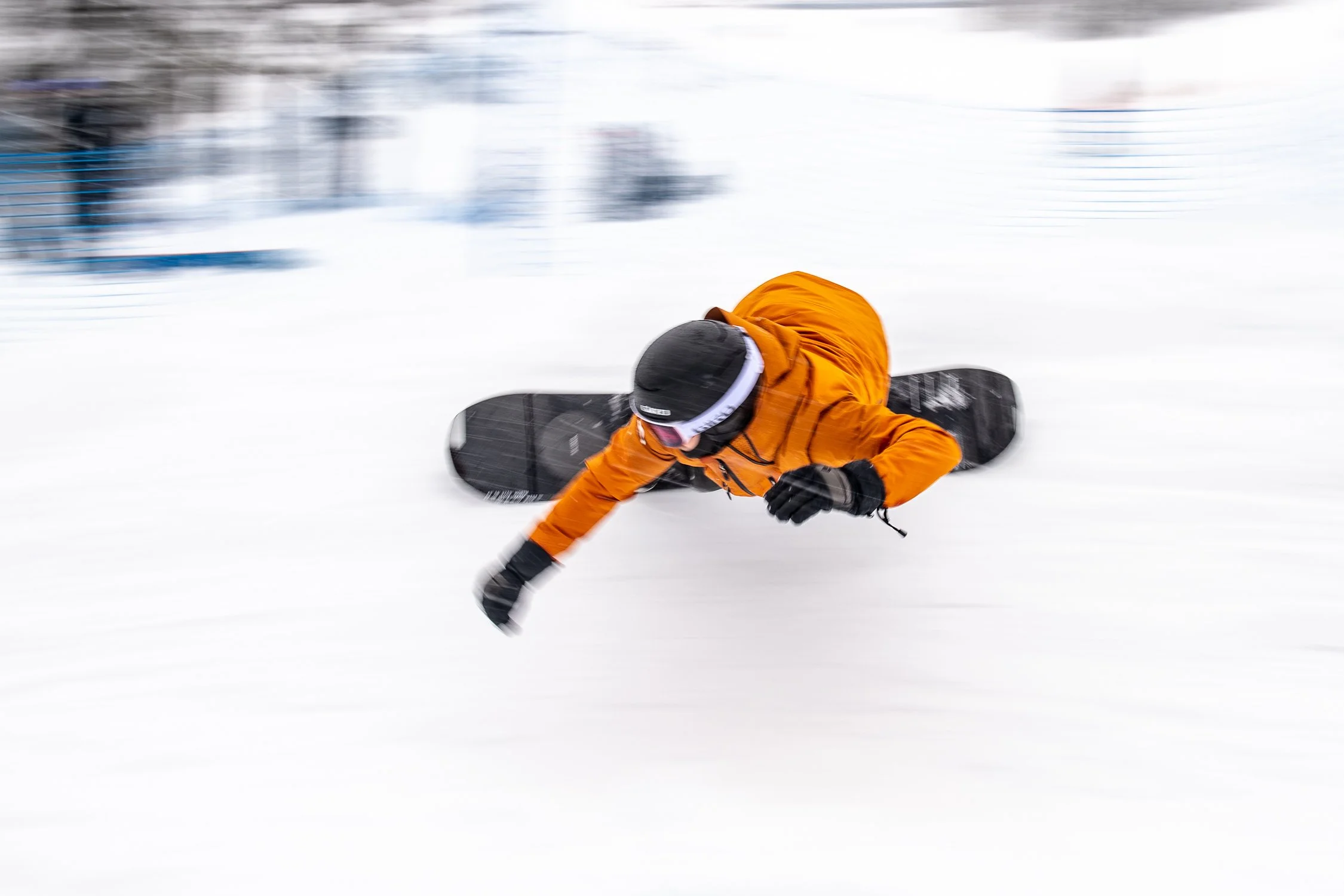 Person in an orange jacket and black helmet falling on snow while snowboarding