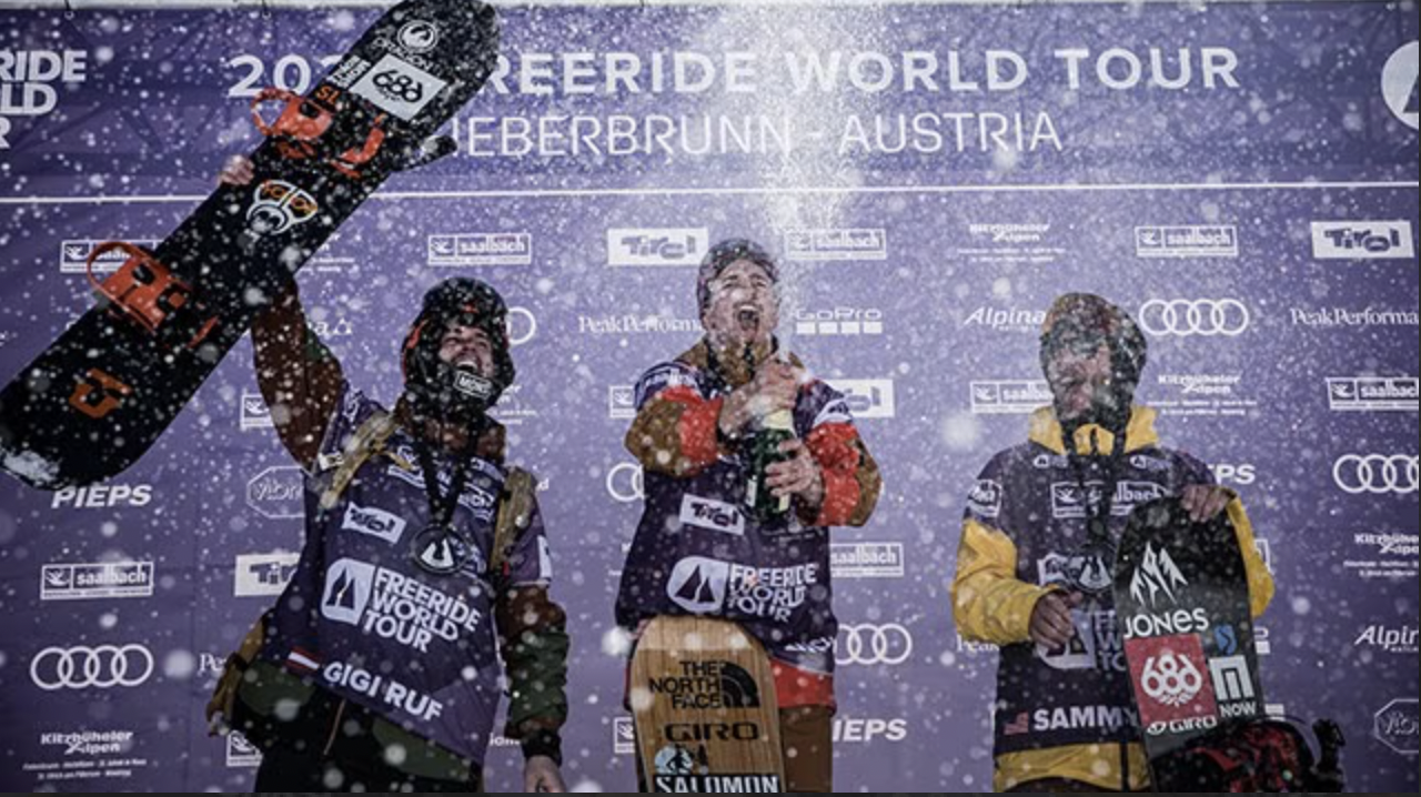 Three male snowboarders on the winners' podium celebrating at the Freeride World Tour event in Leebrun, Austria, with snow falling around them.