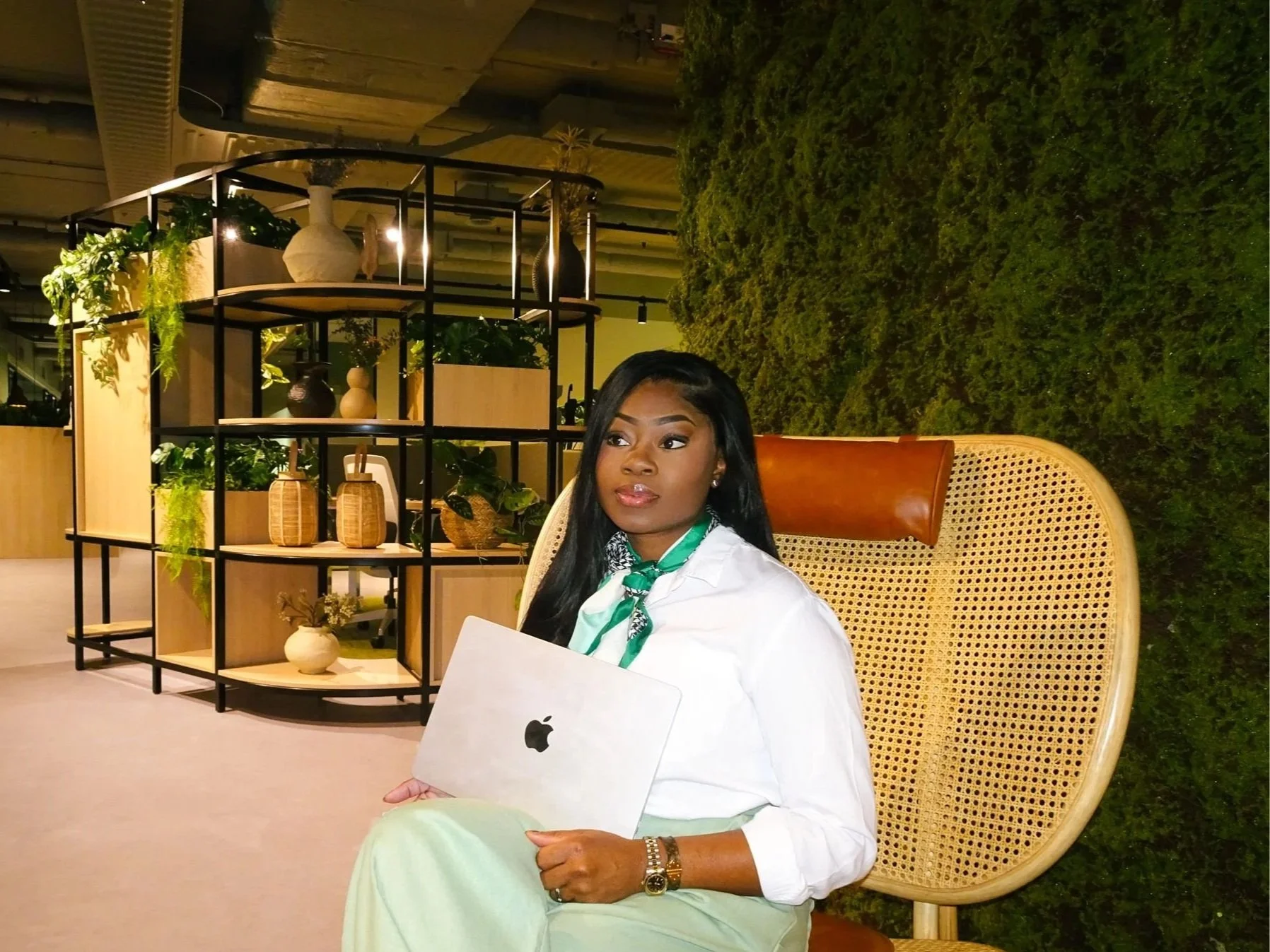 Een vrouw zit op een grote houten stoel met een rieten bekleding, ze houdt een MacBook in haar hand en kijkt naar rechts. Ze draagt een witte blouse, een groene sjaal en heeft lange zwarte haar. De achtergrond bestaat uit een groene muur en een witte houten scheidingswand met decoratieve vazen en planten.