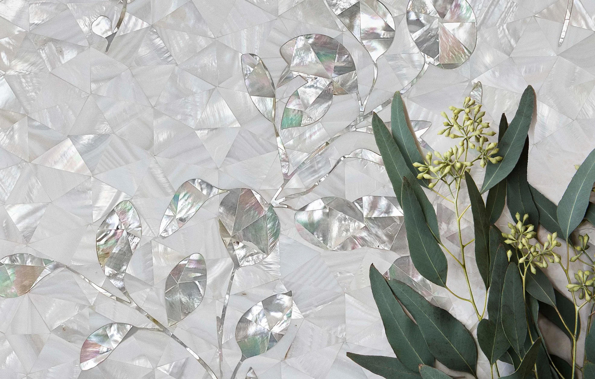 A decorative background with iridescent, symmetry-shaded, mother-of-pearl-like tiles and a sprig of green eucalyptus leaves with small buds.