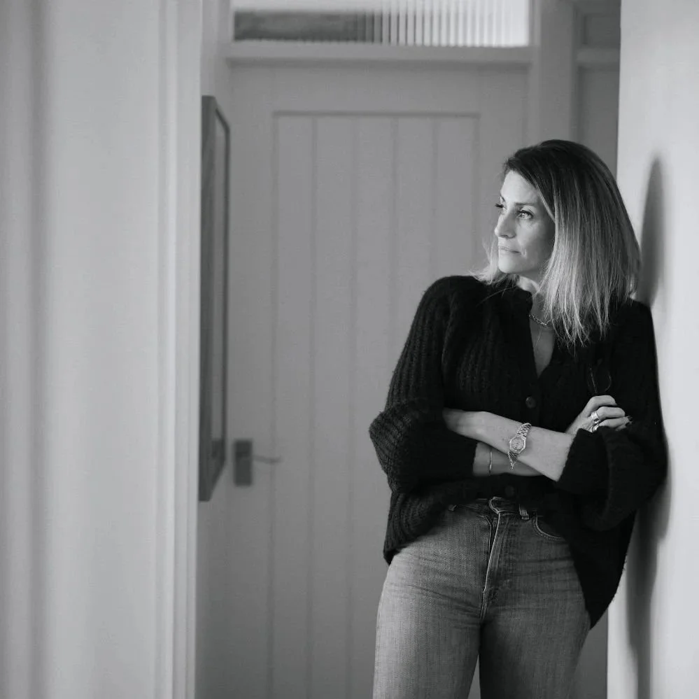 A woman with shoulder-length hair in a black sweater and jeans, leaning against a white wall, looking thoughtfully to the side in a black-and-white photograph.