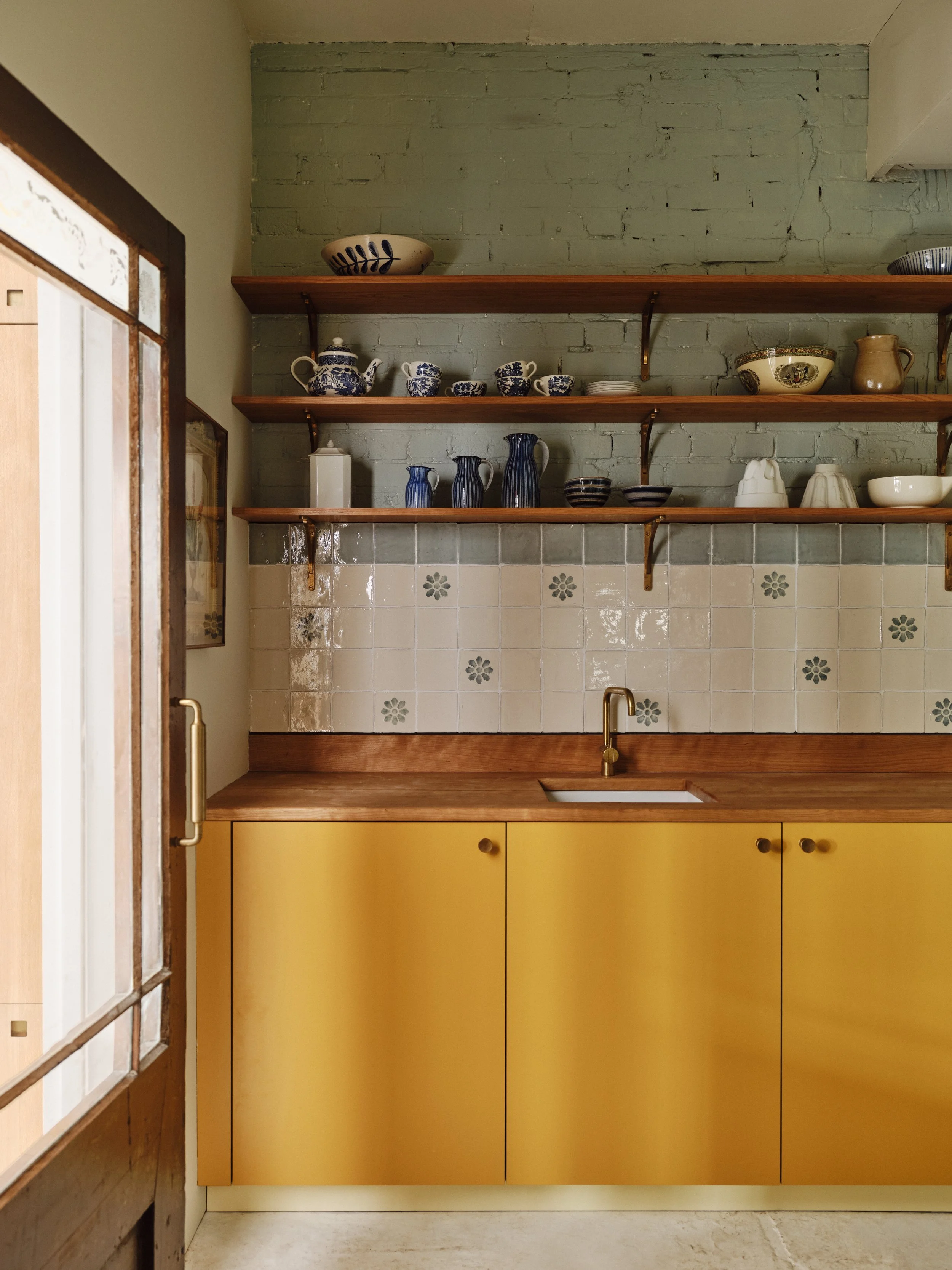A vintage kitchen with yellow cabinets, a wooden countertop, a small sink, and open wooden shelves with blue and white dishware, against a green brick wall.