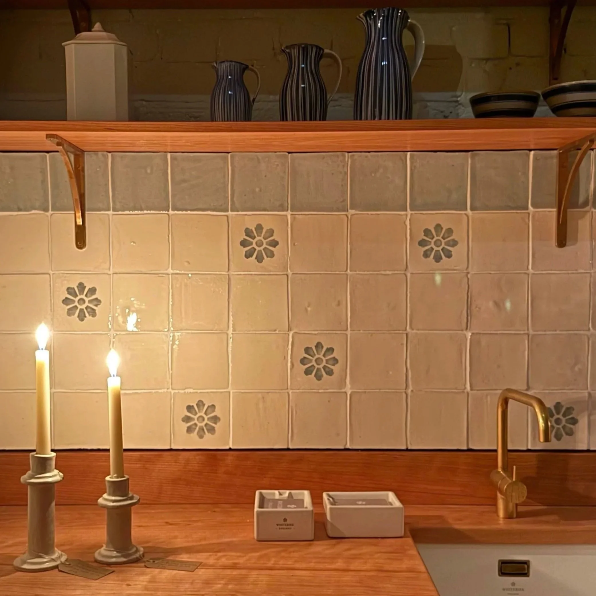 A kitchen scene with a tiled backsplash featuring beige tiles with dark flower patterns, a wooden countertop with two white candles in candle holders, a small sink with a gold faucet, and decorative black vases and bowls on a top shelf.