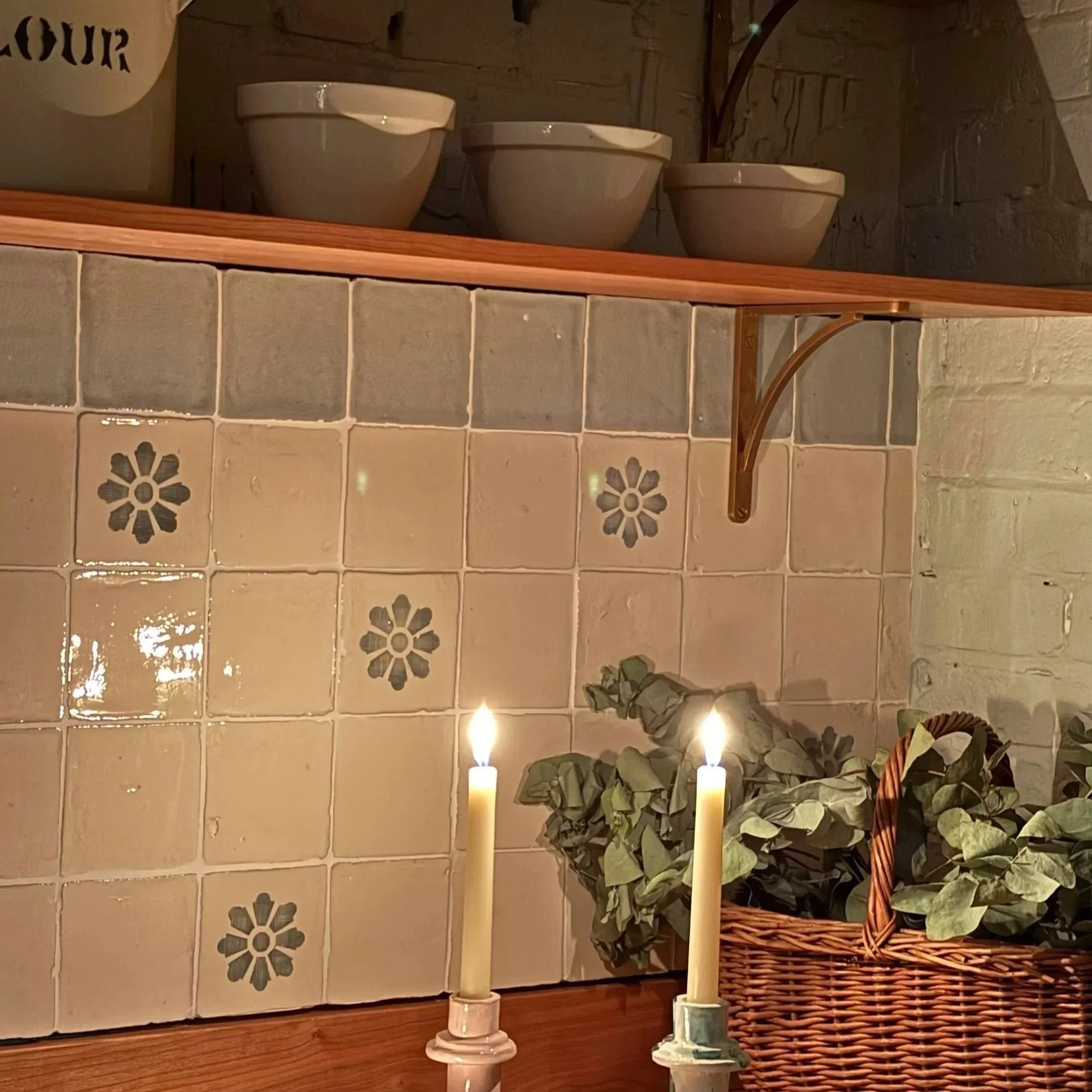 Two lit candles in candle holders, a woven basket with green leafy plants, and a tiled wall with decorative flowers in a cozy indoor setting.