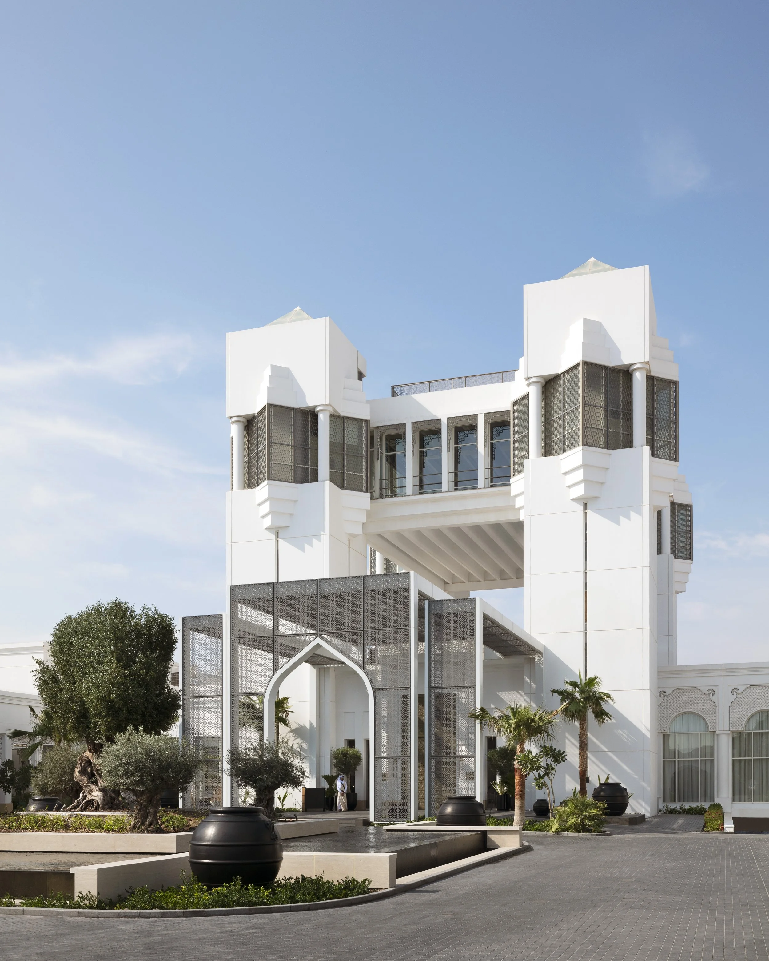 Modern white building with Arabic-inspired entrance, palm trees, and potted plants in a landscaped area.