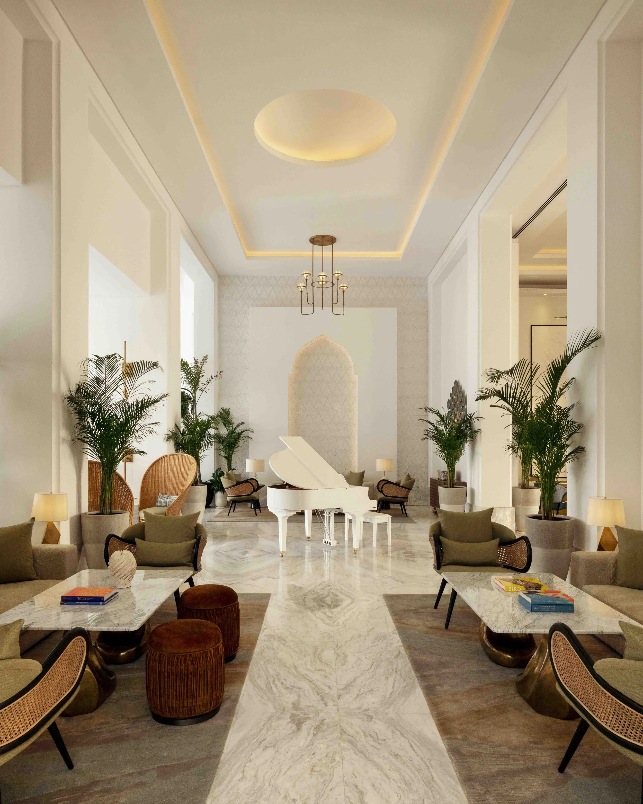Elegant hotel lounge with a white grand piano at the center, surrounded by cushioned chairs and sofas, large potted plants, marble floors, and a high ceiling with integrated lighting.