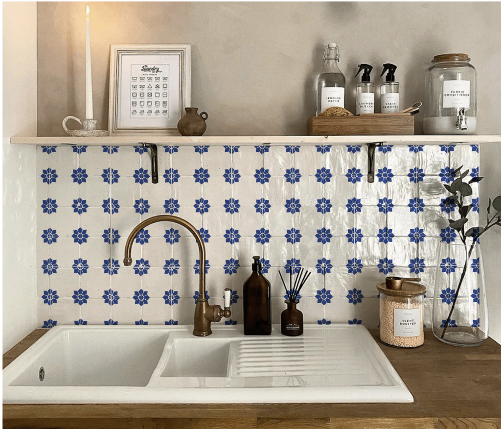 Kitchen sink area with a white double basin sink and bronze faucet. Behind the sink, there's a blue and white patterned tile backsplash. On the wooden countertop, there are brown soap dispensers, a jar labeled 'Scent Booster,' and a tall glass vase w