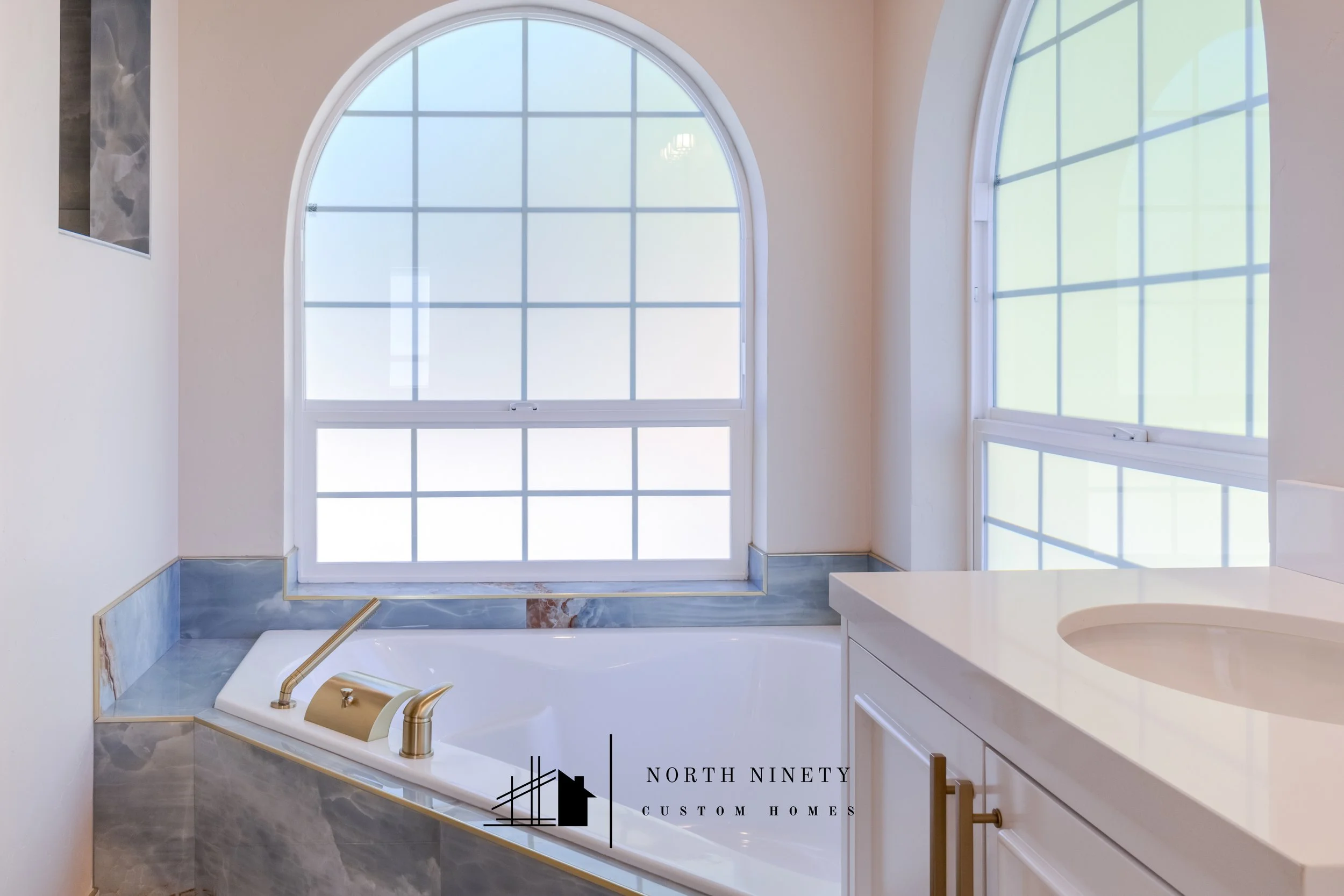 A bathroom with a large arched window, a corner bathtub with gold fixtures, white walls, and a marble base. There is a vanity with a white countertop and a round sink. North Ninety Custom Homes | Treasure Valley | Idaho