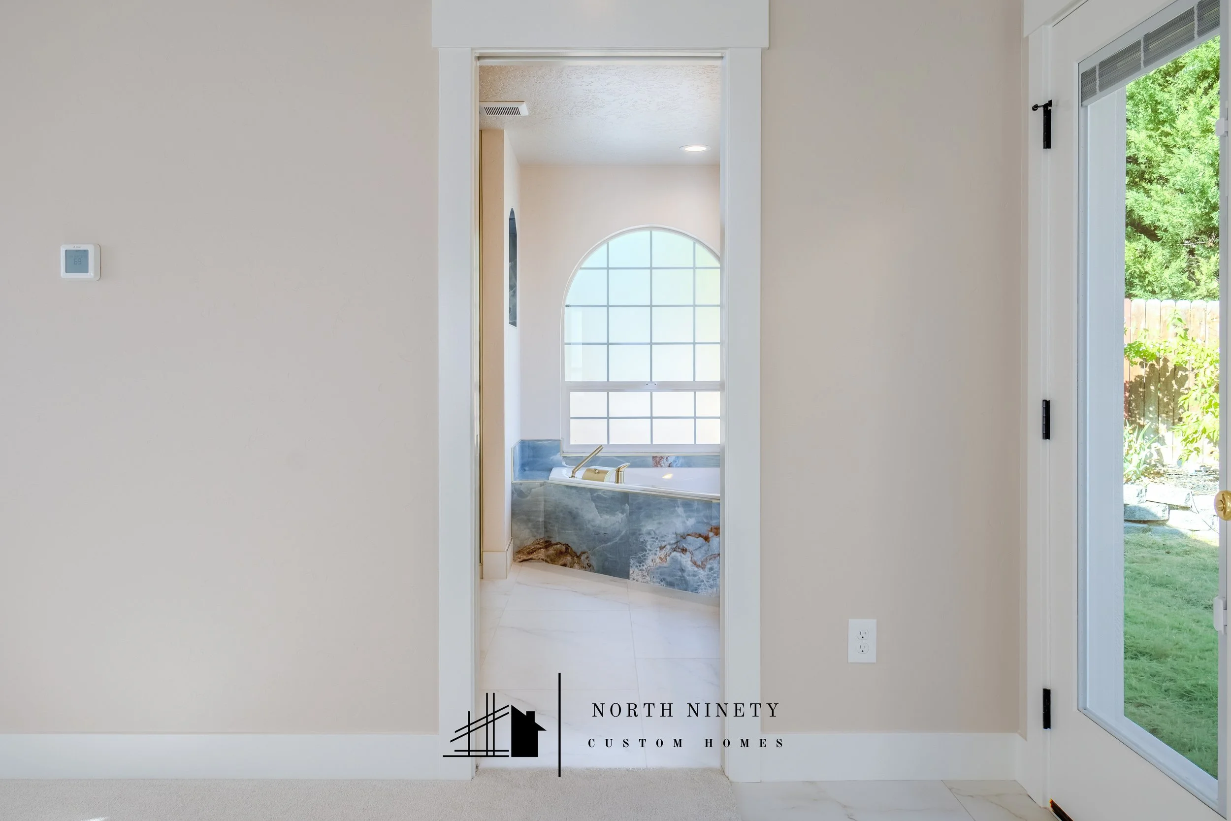 View from inside a house looking into a bathroom with a large arched window, bathtub, and marble accents, with a sliding glass door to a backyard visible on the right. North Ninety Custom Homes | Treasure Valley | Idaho