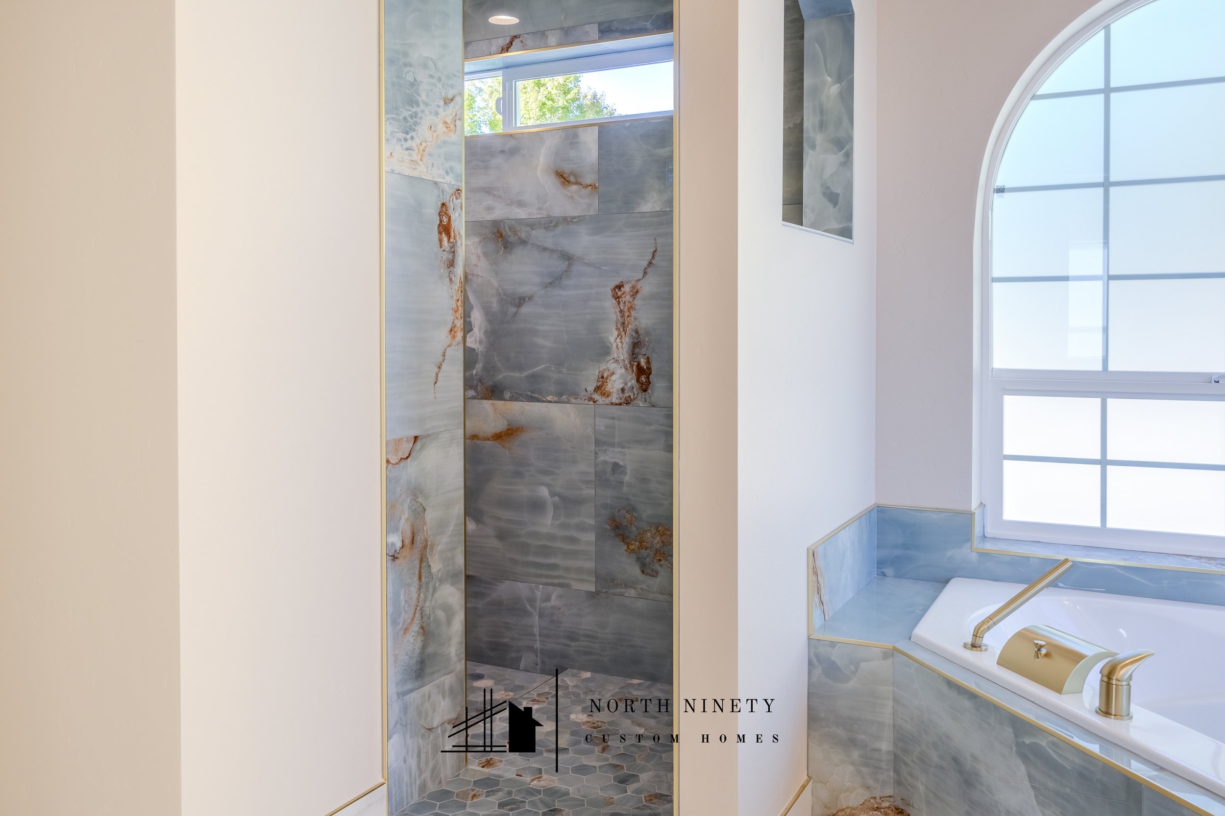 Interior view of a bathroom with large marble tiles, a vanity with gold fixtures, and a large arched window allowing natural light. North Ninety Custom Homes | Treasure Valley | Idaho