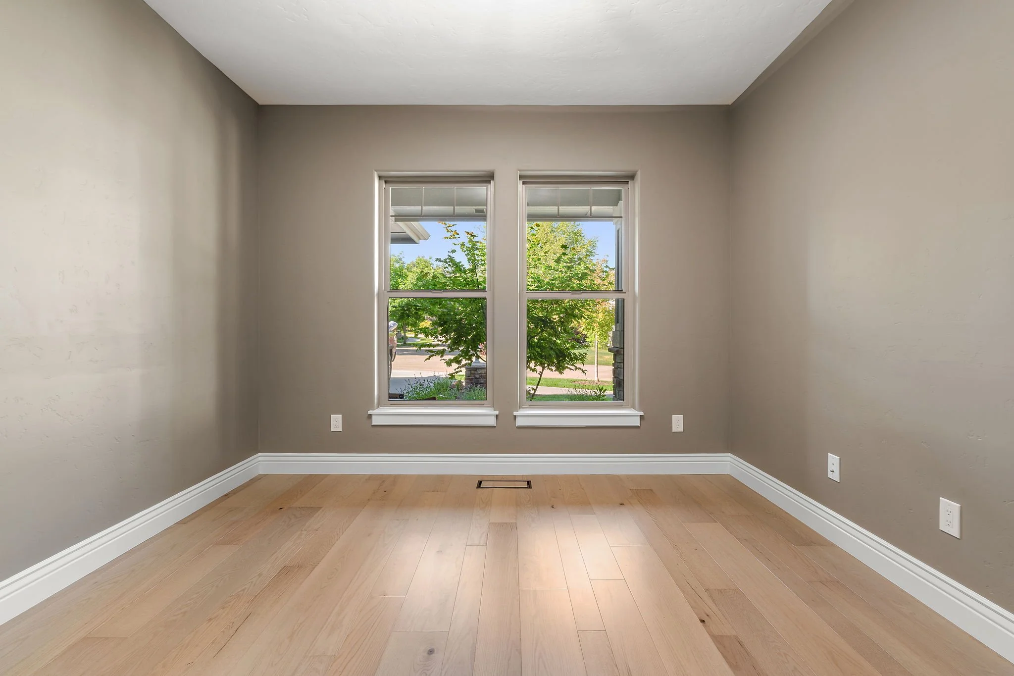 Empty room with beige walls, two windows showing a green tree outside, light wood flooring, and white trim and baseboards. North Ninety Custom Homes | Treasure Valley | Idaho