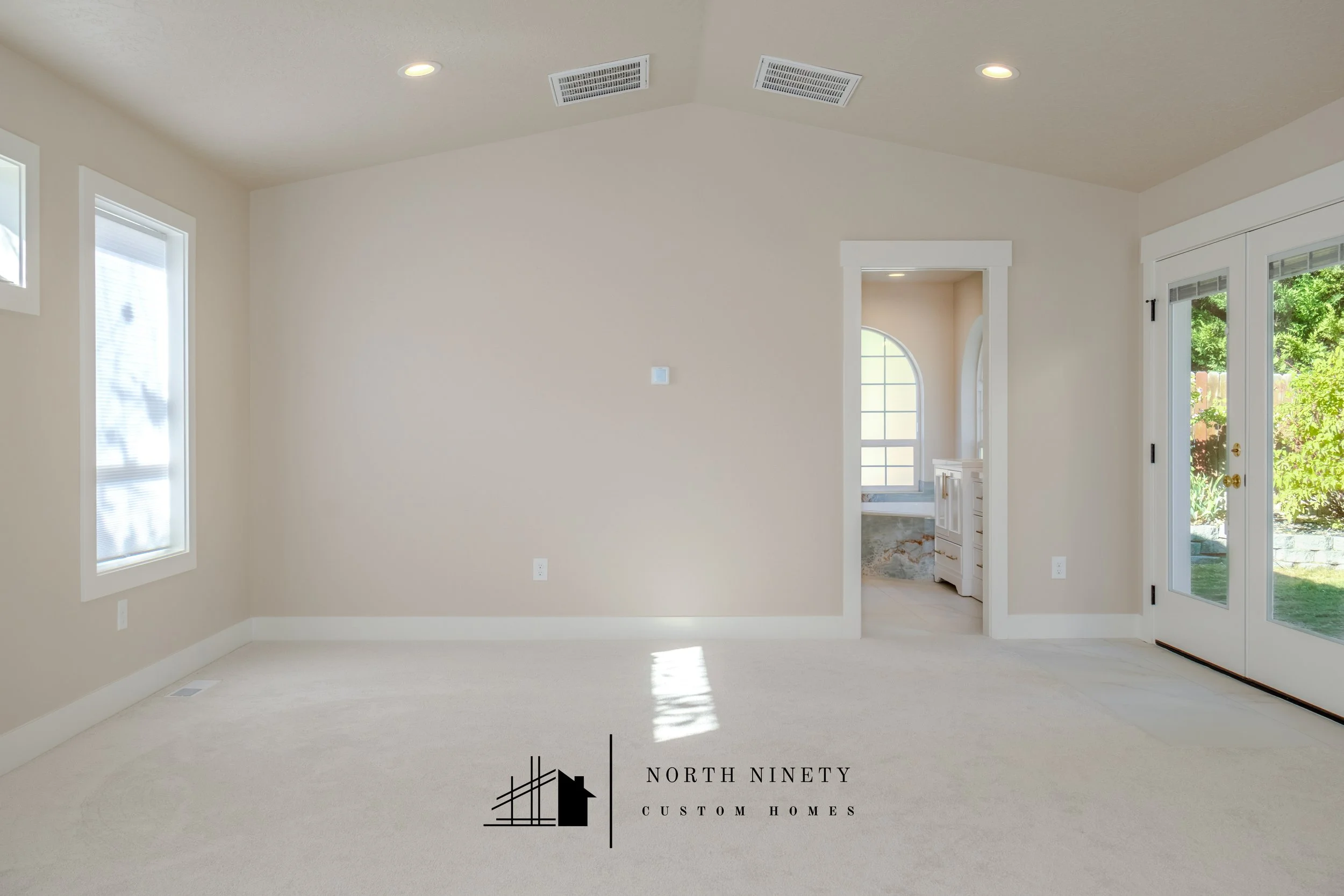 Empty living room with beige walls, white trim, natural light from windows, and an open doorway leading to another room with a window. North Ninety Custom Homes | Treasure Valley | Idaho