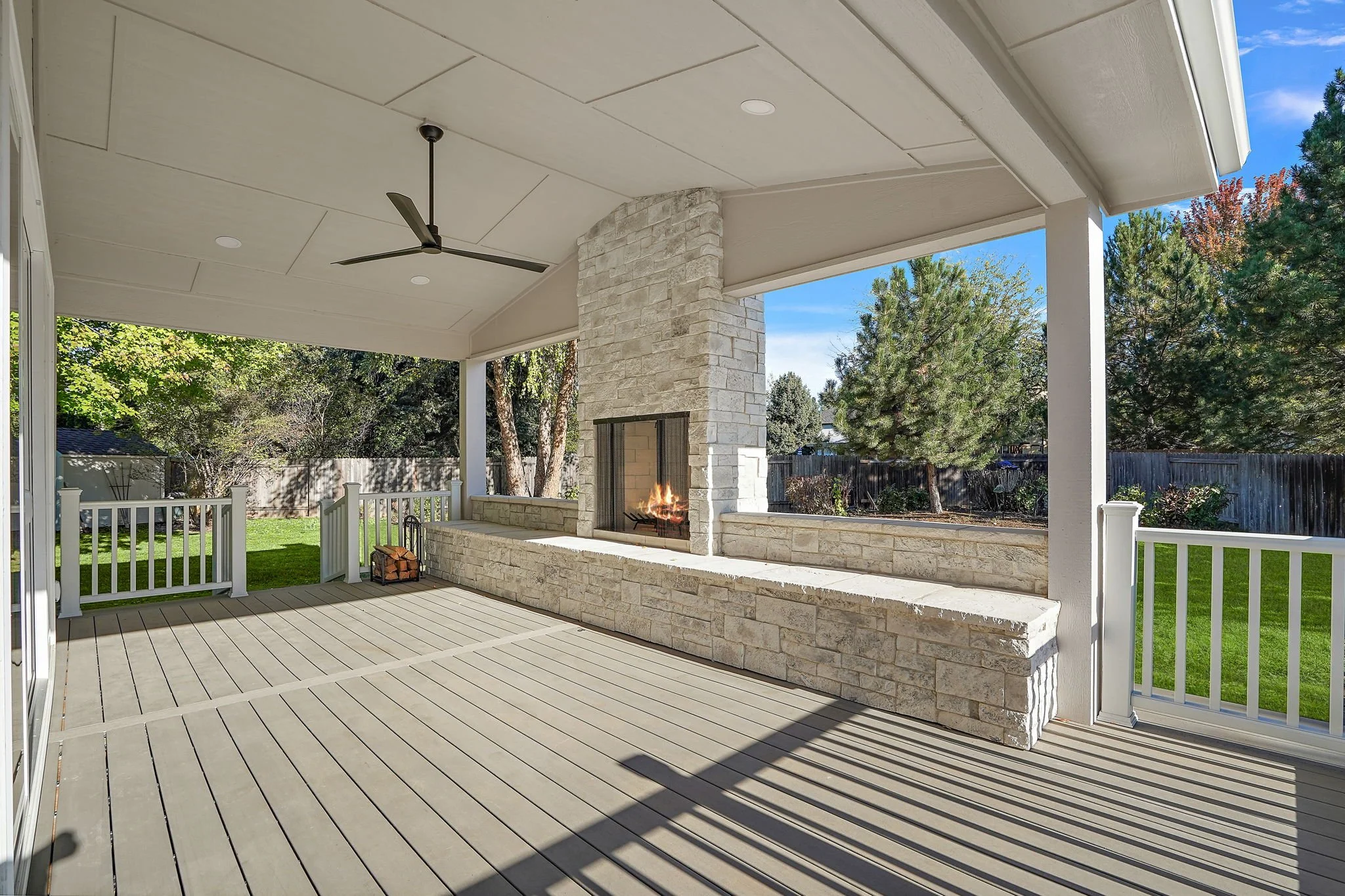 Covered backyard patio with a fire feature, ceiling fan, and a view of a green lawn and trees.