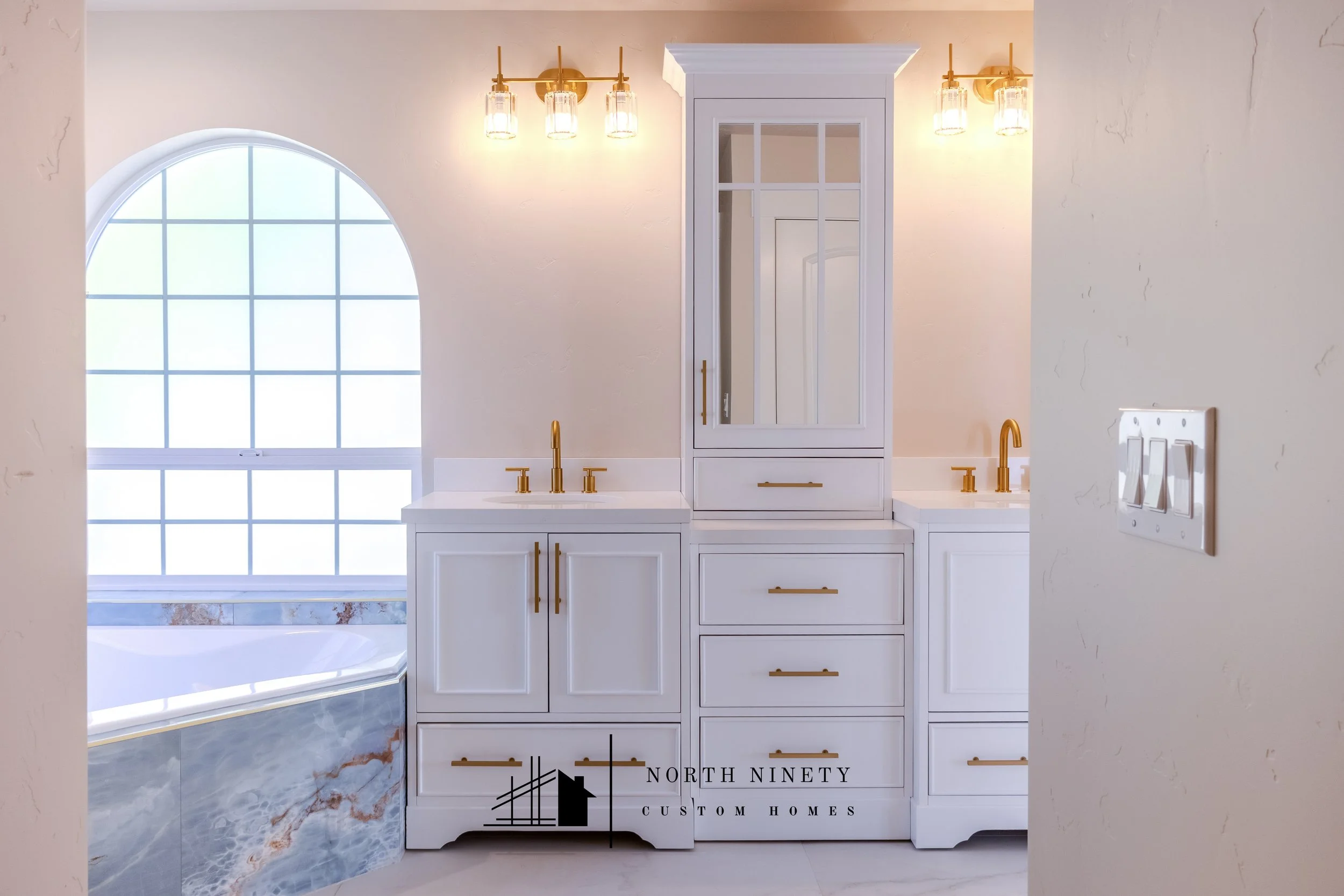 Modern bathroom with white cabinetry, gold hardware, and large window with grid design. Features a bathtub with marble-style tiles and two wall-mounted light fixtures with glass shades. North Ninety Custom Homes | Treasure Valley | Idaho
