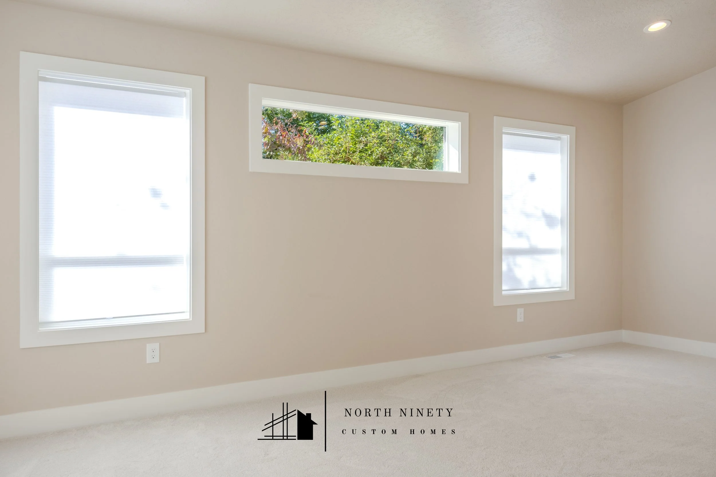 Empty room with three windows, two vertical and one horizontal, showing greenery outside, beige walls, carpeted floor, recessed ceiling light, and an outlet near the floor. North Ninety Custom Homes | Treasure Valley | Idaho