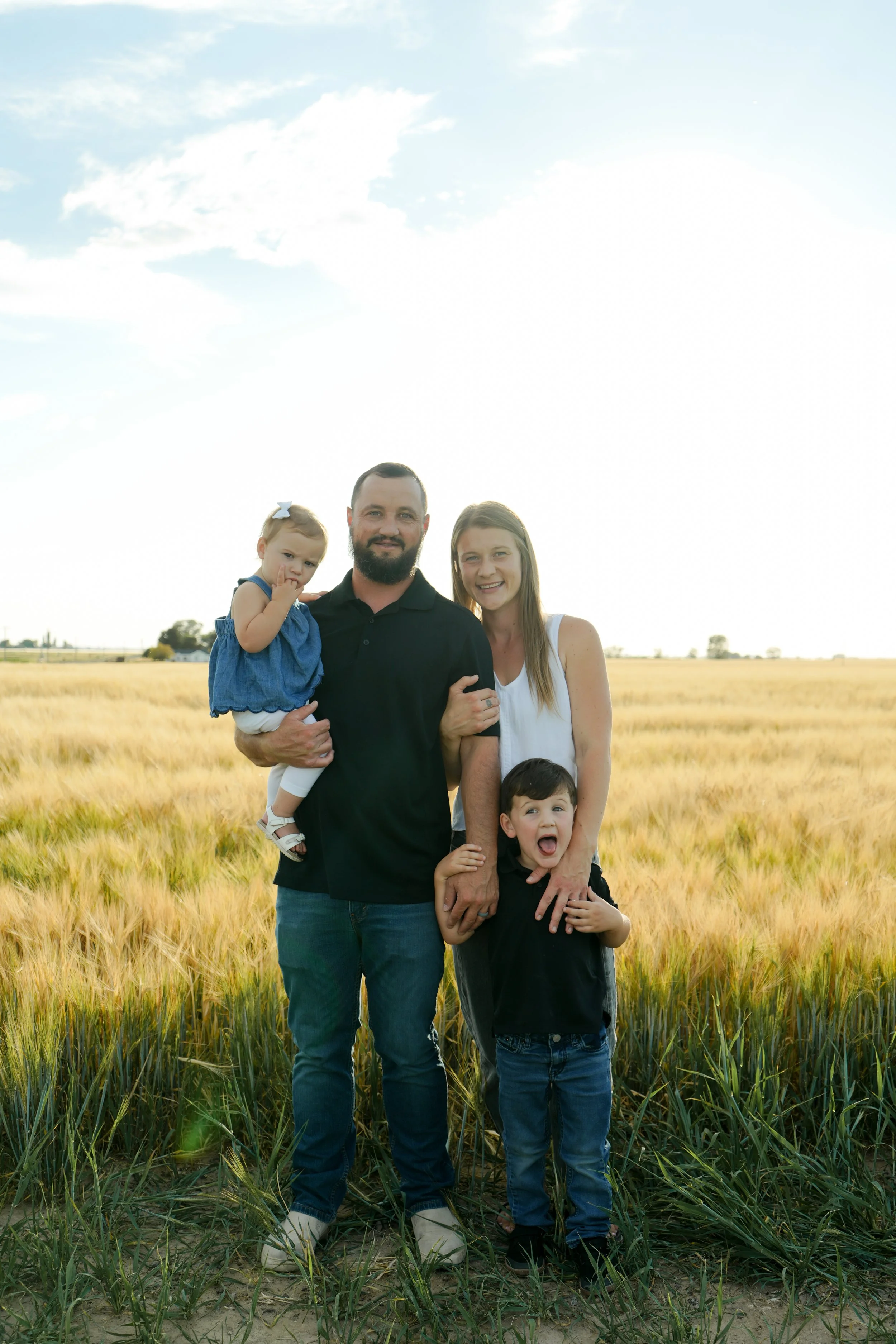 A family of five standing in a field of tall grass, smiling, with a farm landscape and partly cloudy sky in the background.