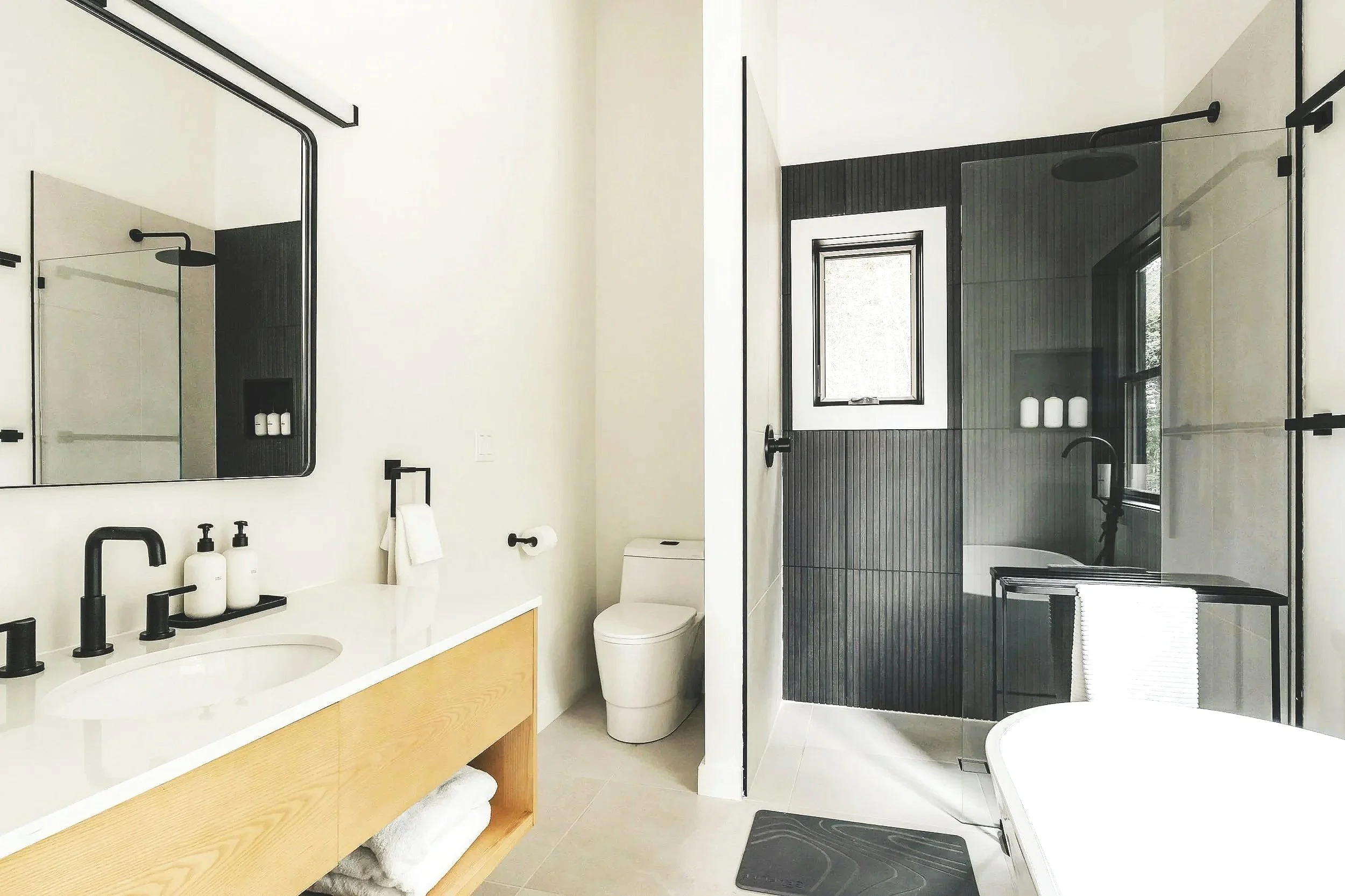 A modern bathroom with a dual-sink vanity, large mirror, black faucets, black towel racks, a toilet, a window, and a glass-enclosed shower with black fixtures and a window, all featuring a minimalist black and white color scheme.