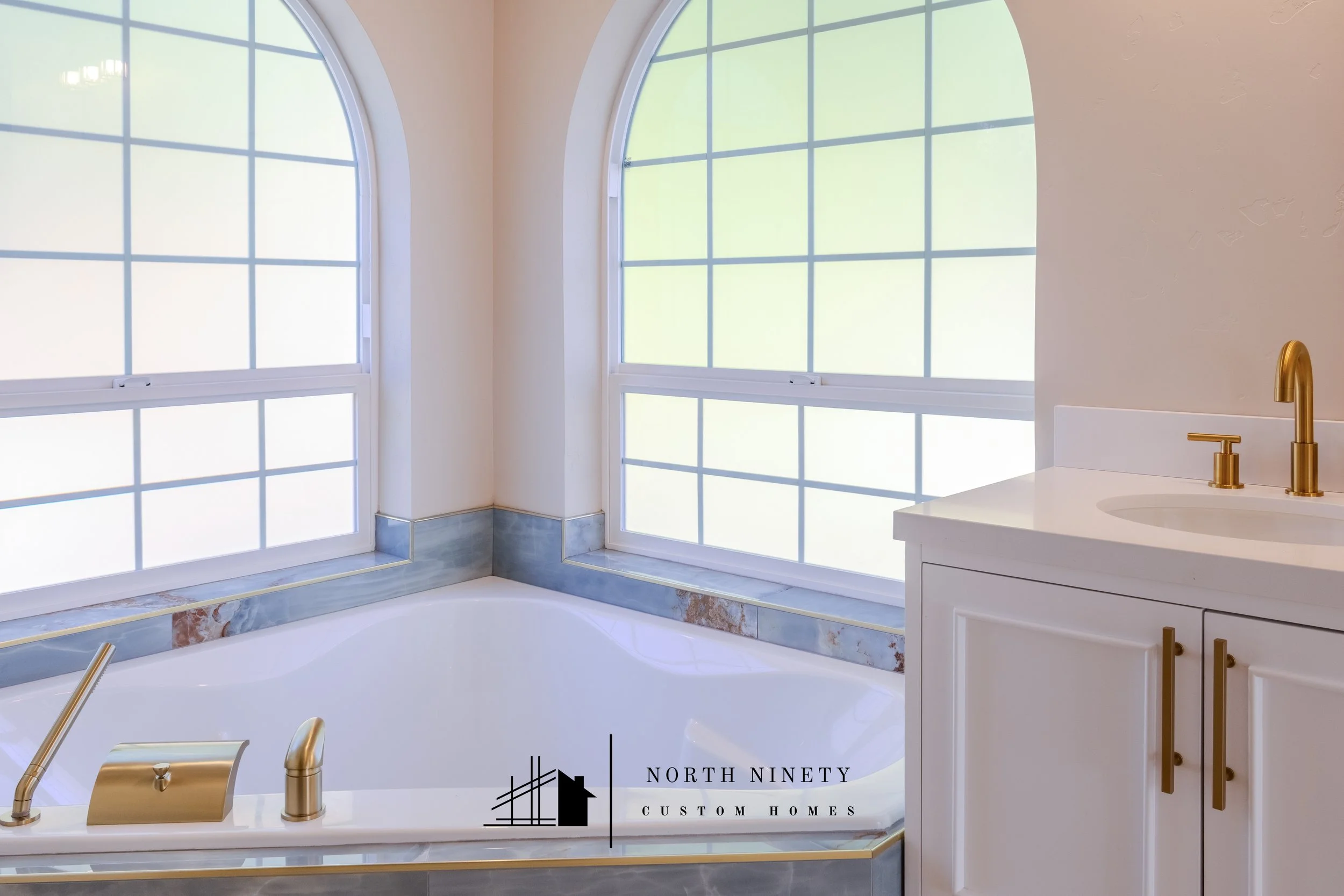 A bathroom with large arched windows, white walls, a bathtub with a marble surround, and a white vanity with gold fixtures. North Ninety Custom Homes | Treasure Valley | Idaho