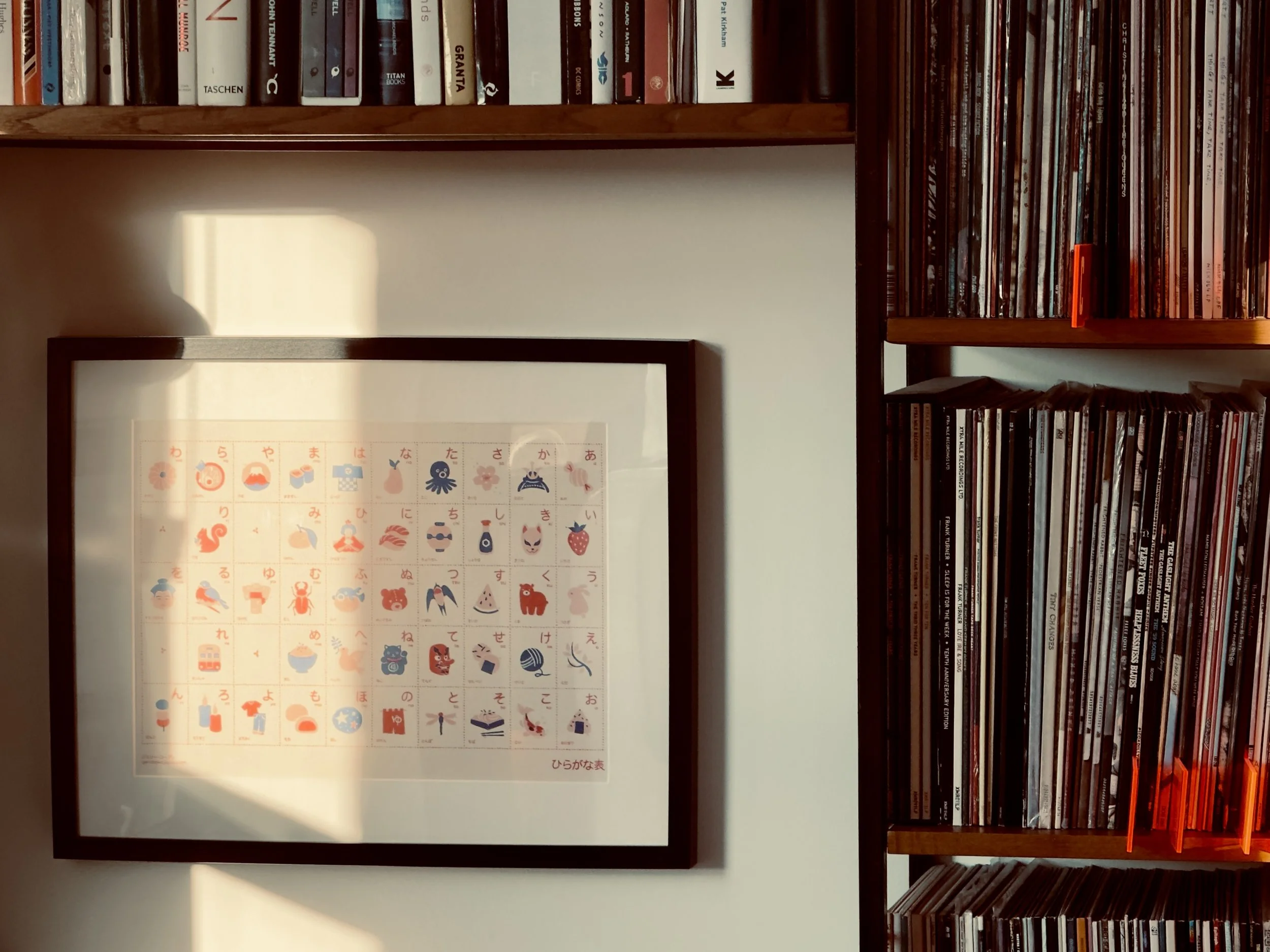 A framed poster of the Japanese alphabet with illustrated characters, hanging on a white wall next to a wooden bookshelf filled with books and vinyl records.