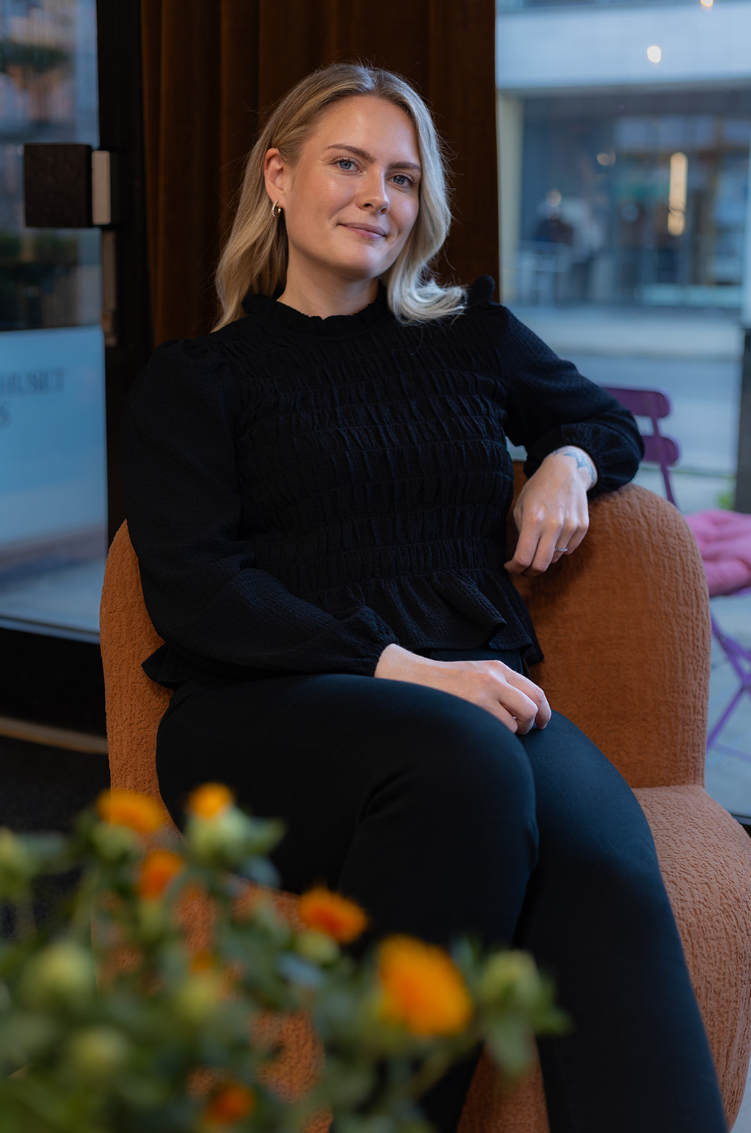 Kvinne i svart genser sitter komfortabelt i en stol i et innendørs miljø.