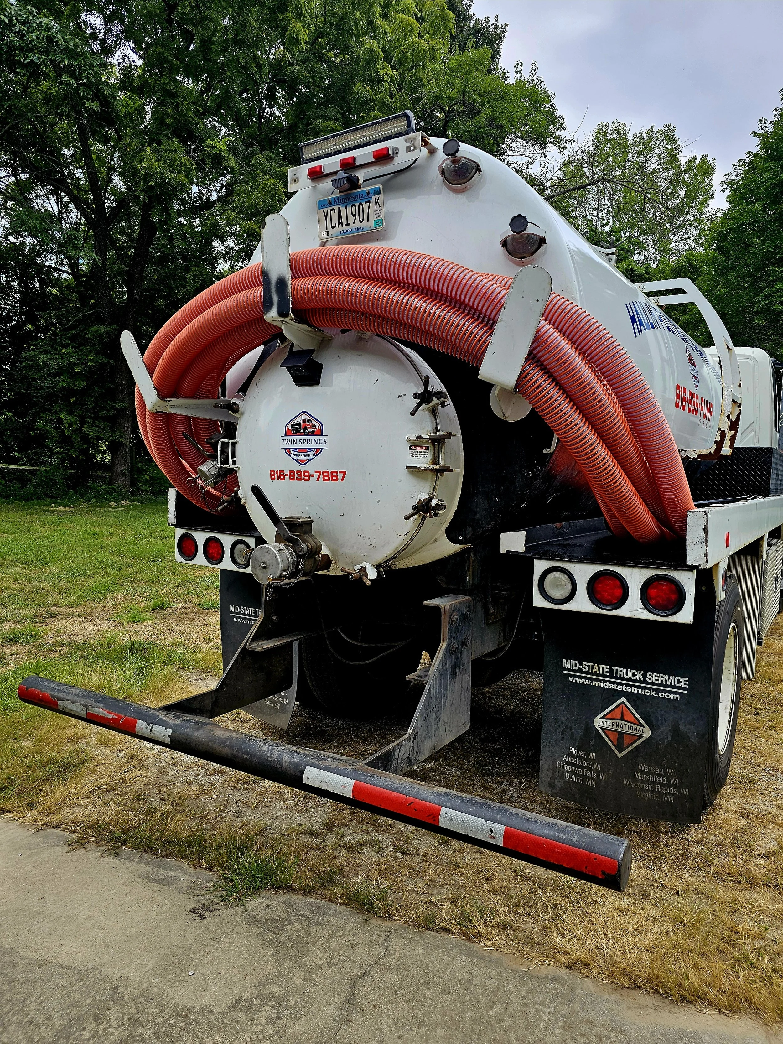 twin springs vacuum truck with orange hoses, parked on grass near trees.