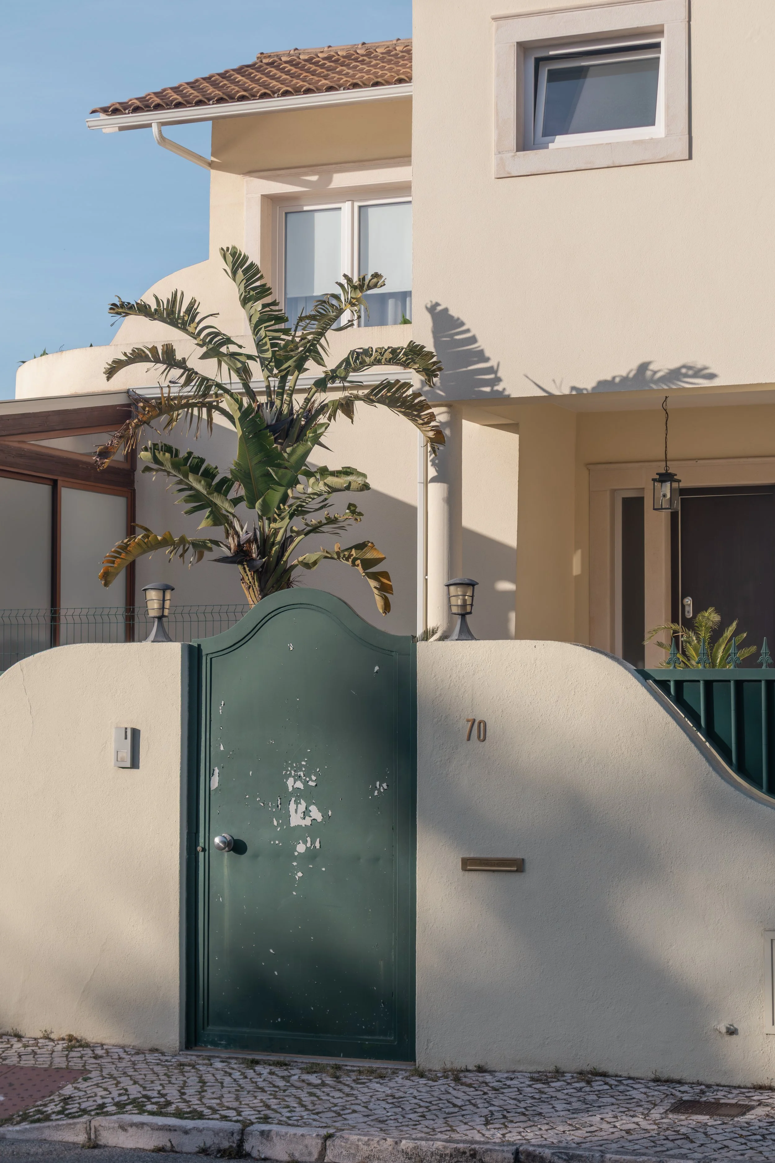A house with a green metal gate, small palm tree, and cream-colored exterior walls with windows. The house number is 70.