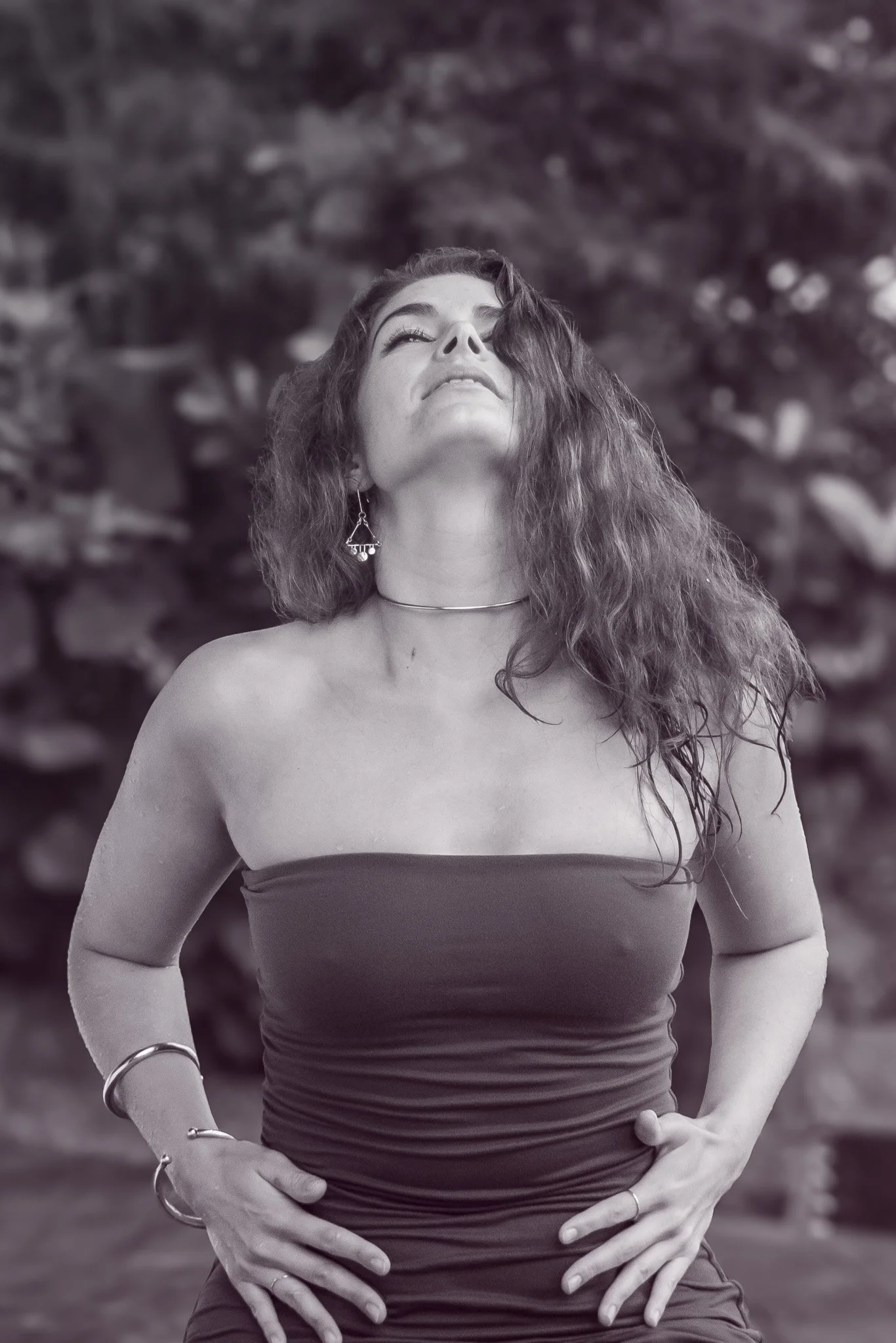 A woman with curly hair and earrings in an outdoor setting, looking upward with her hands on her hips in a strapless top.