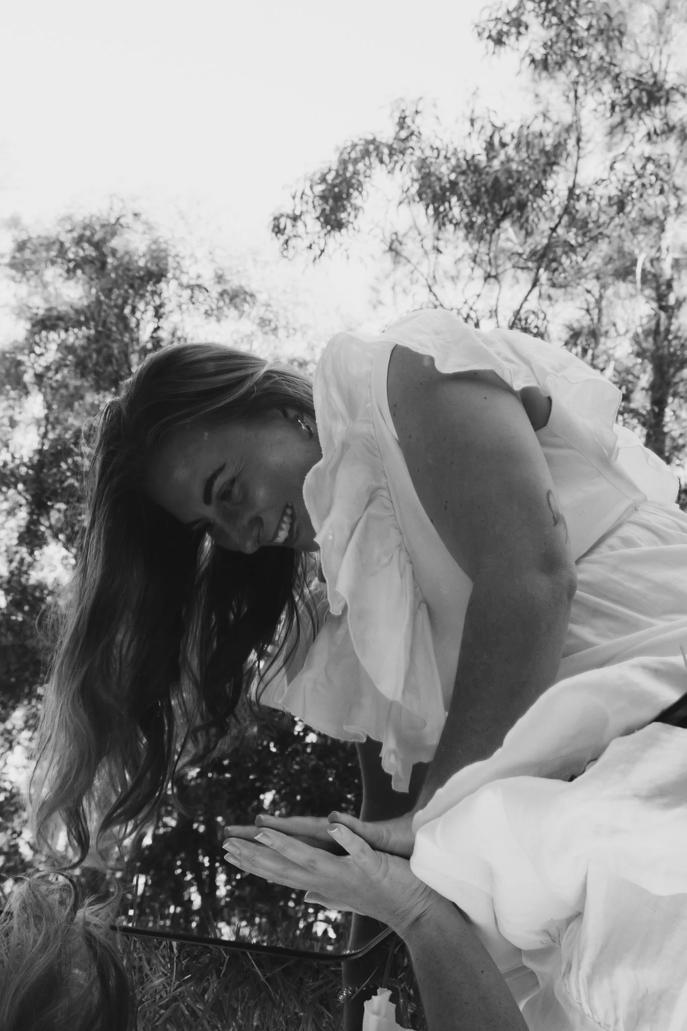 A woman with long hair smiling and leaning over a mirror outdoors, with trees and sky in the background, in black and white.