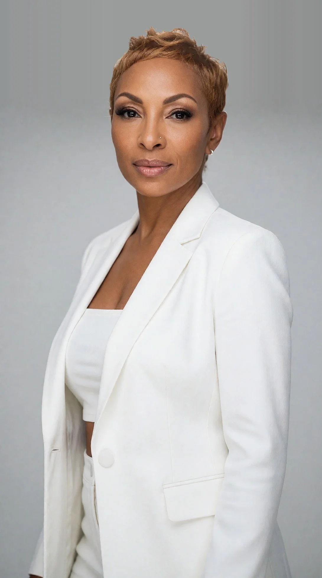 Portrait of a woman with short, styled hair, dressed in a white blazer and top, against a plain gray background.