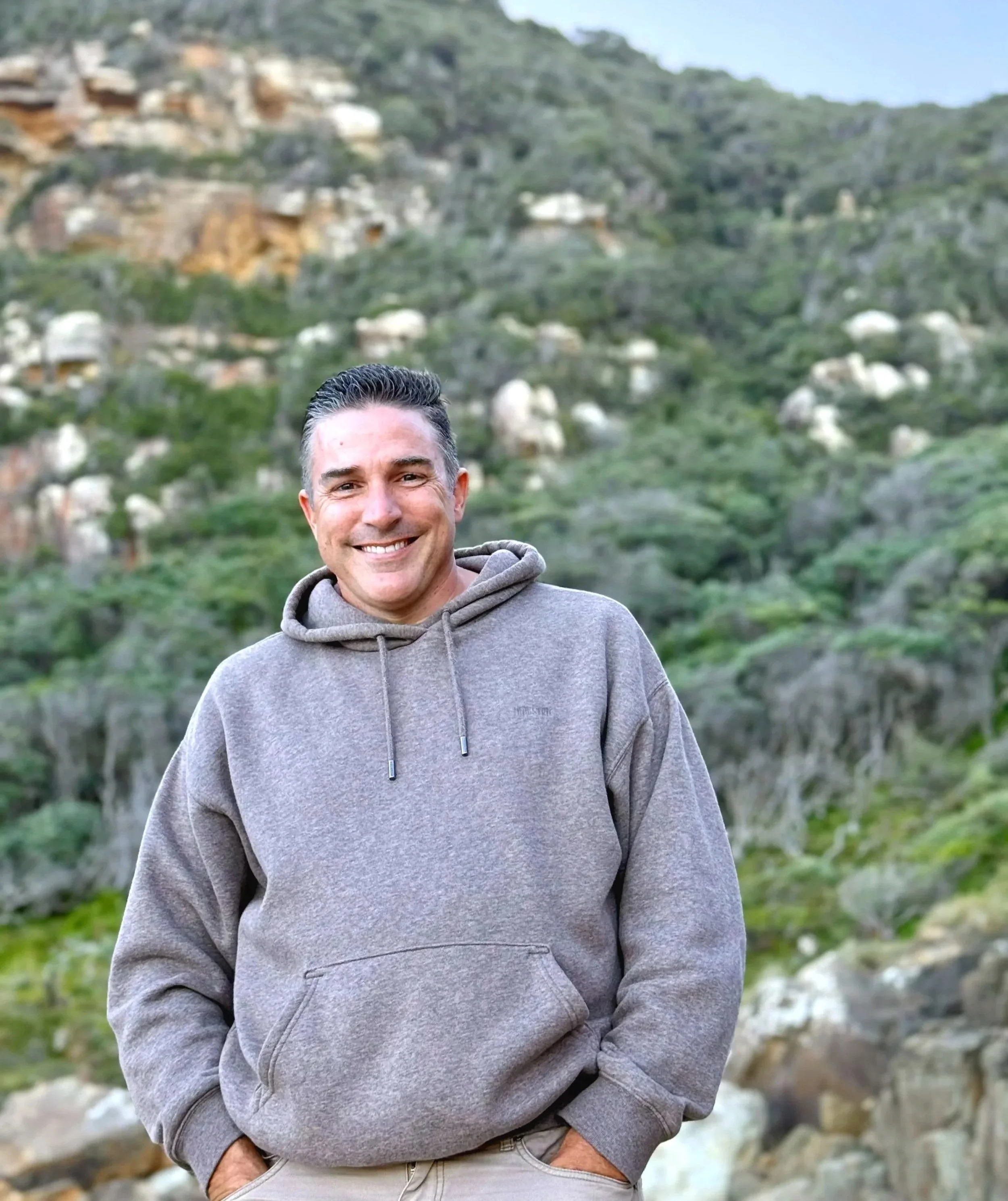 Justin smiling outdoors in front of a rocky, green hillside. - Jason's bio, The Inner Shift Ekklesia