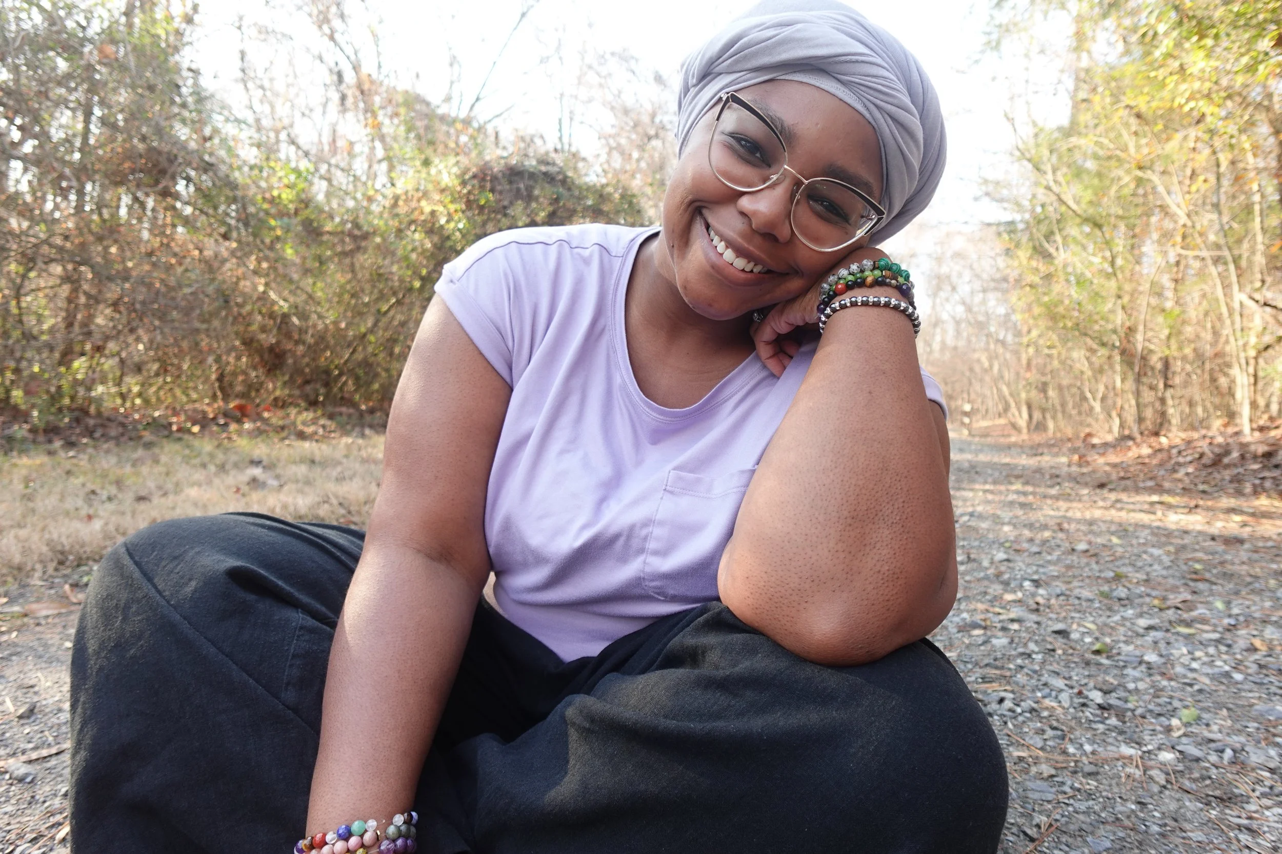 Portrait of a woman with glasses, smiling, wearing a yellow blazer and white top, outdoors with green trees in the background.