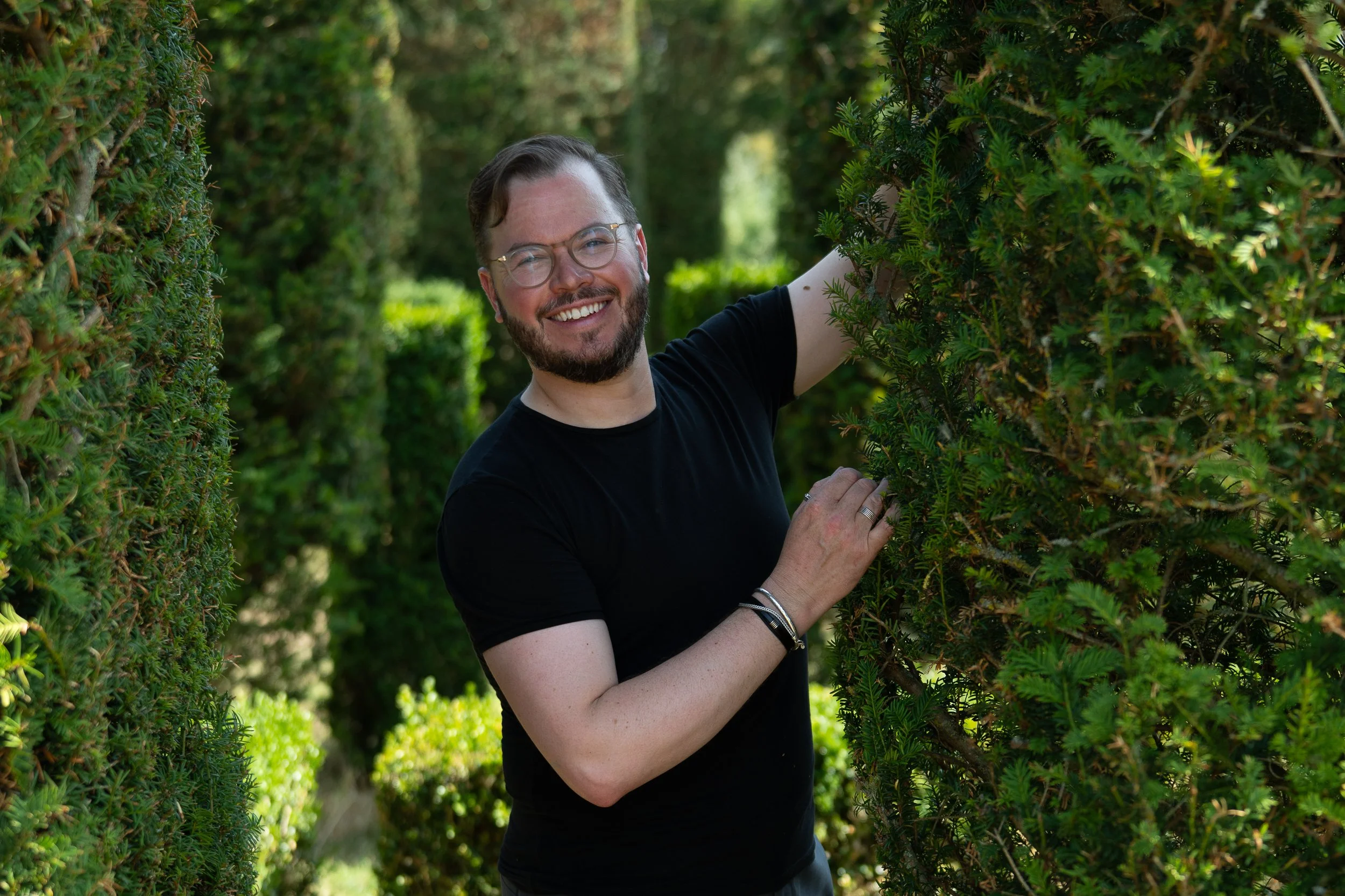 A man with glasses and a beard smiling while standing in a hedge maze, surrounded by green bushes and trees.