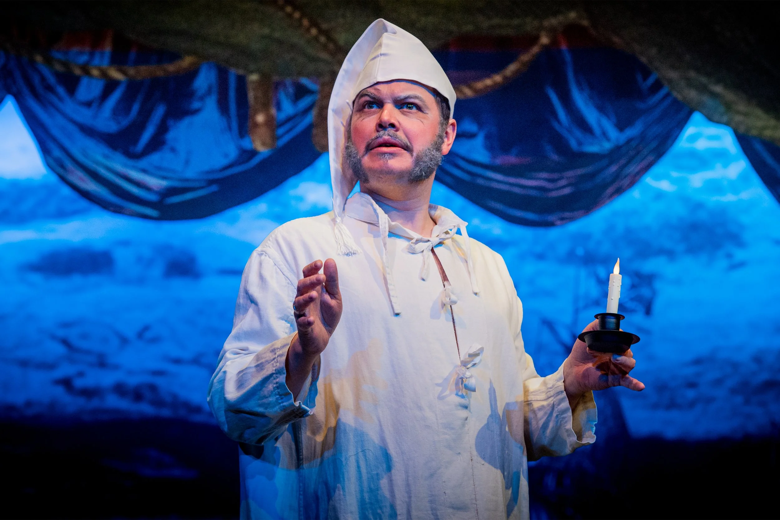 A man dressed in colonial attire, including a white shirt with a tied collar and a white cap, holds a black candlestick with a lit candle. He is on a stage with a backdrop resembling a cloudy blue sky and dark blue drapery overhead.