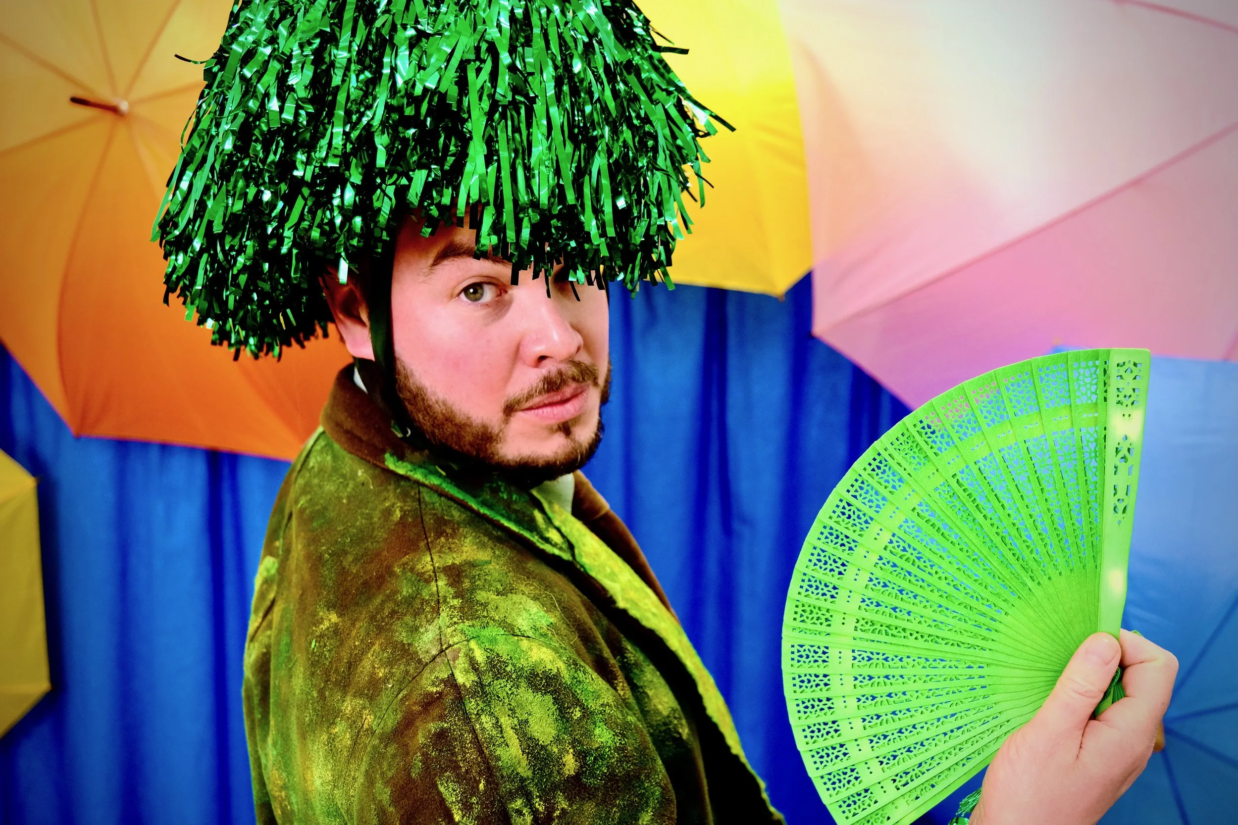 A man holding a green fan and wearing a green tinsel hat, standing in front of colorful umbrellas.