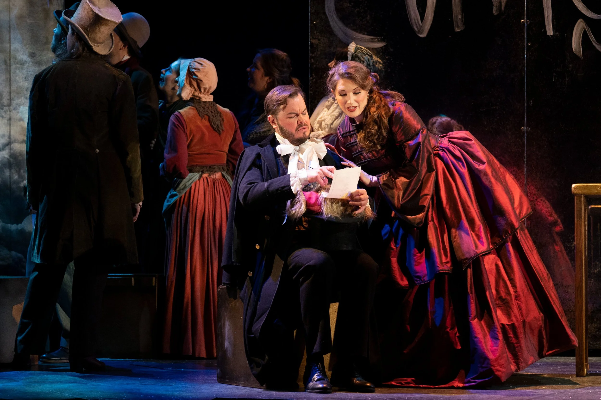 Actors in historical costumes performing on stage, with one seated man reading a paper and a standing woman leaning over him, surrounded by other actors in period clothing.