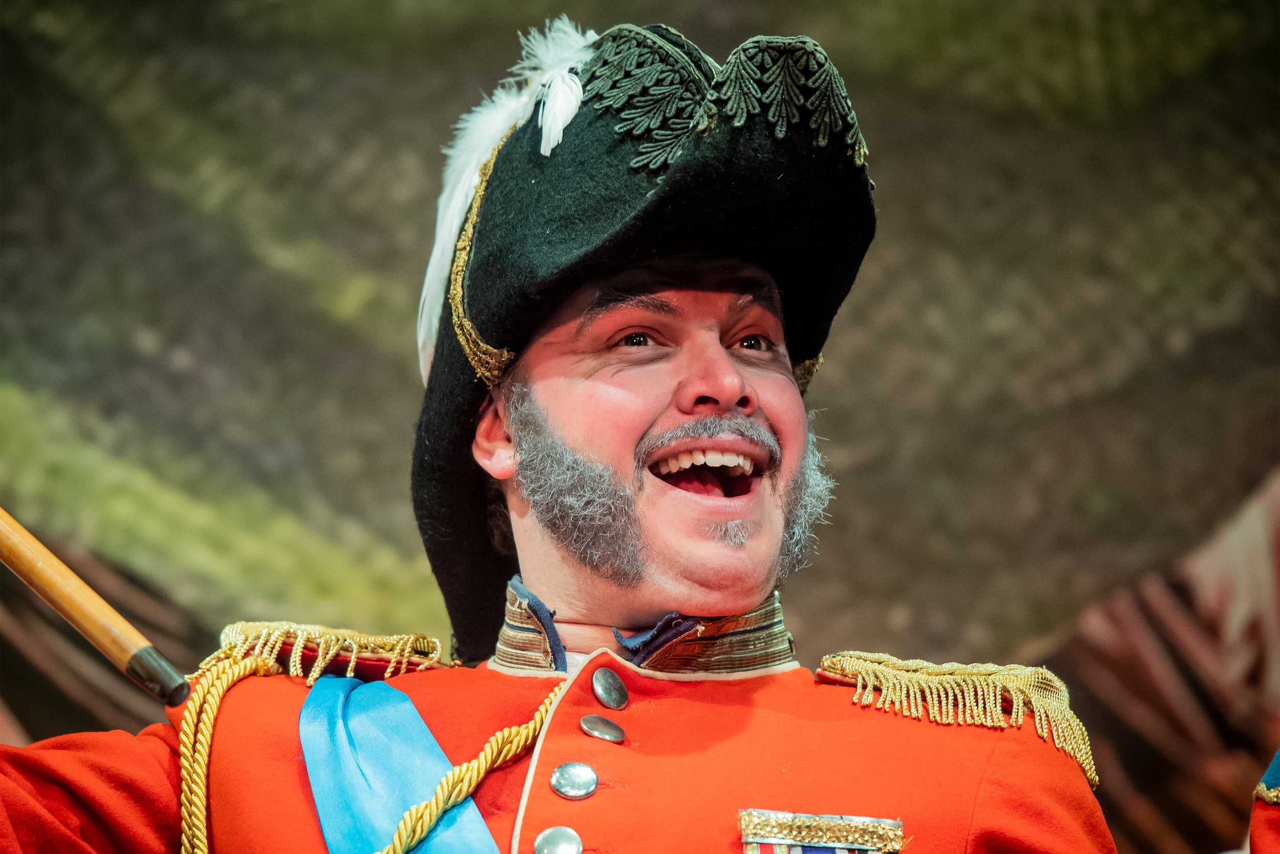 A man dressed as a British soldier with a red coat, gold trim, and Tall hat, smiling broadly. He has a gray beard and mustache, and a background resembling a textured green wall.