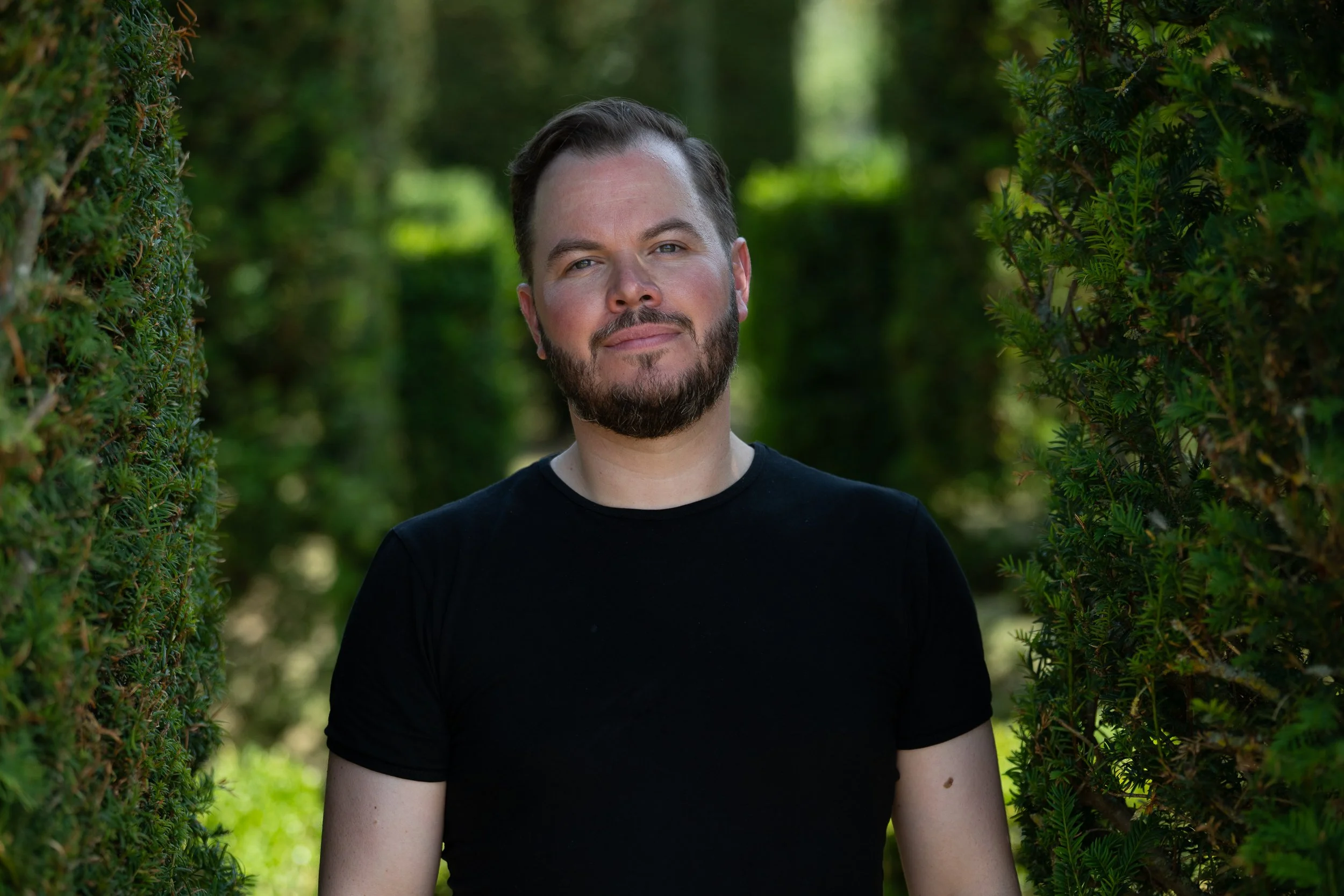 A man with a beard and short brown hair stands outdoors between two green hedges, wearing a black T-shirt.