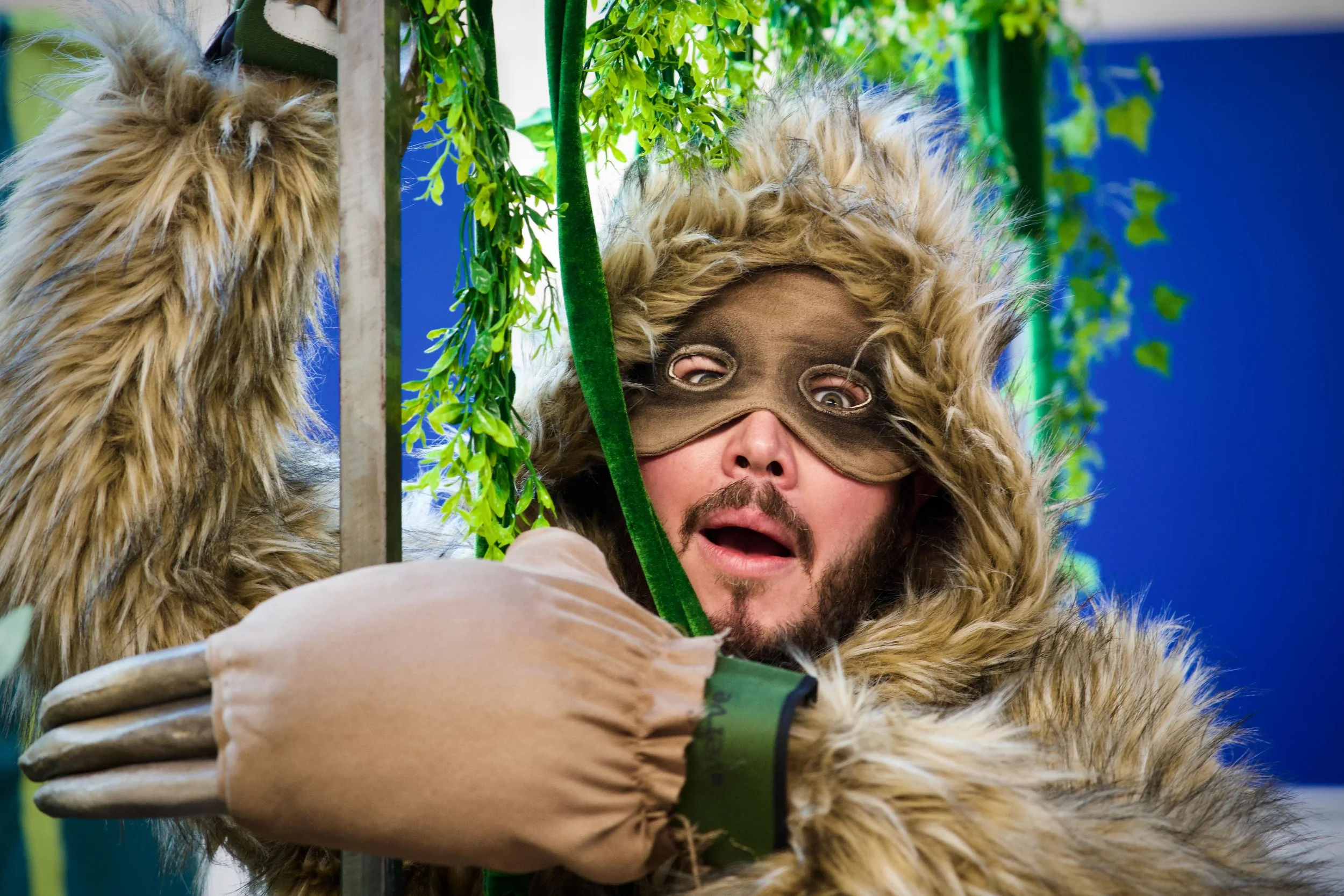Person dressed as a furry animal with a brown mask and furry costume, holding onto a green vine and looking surprised or scared, with greenery and blue background.