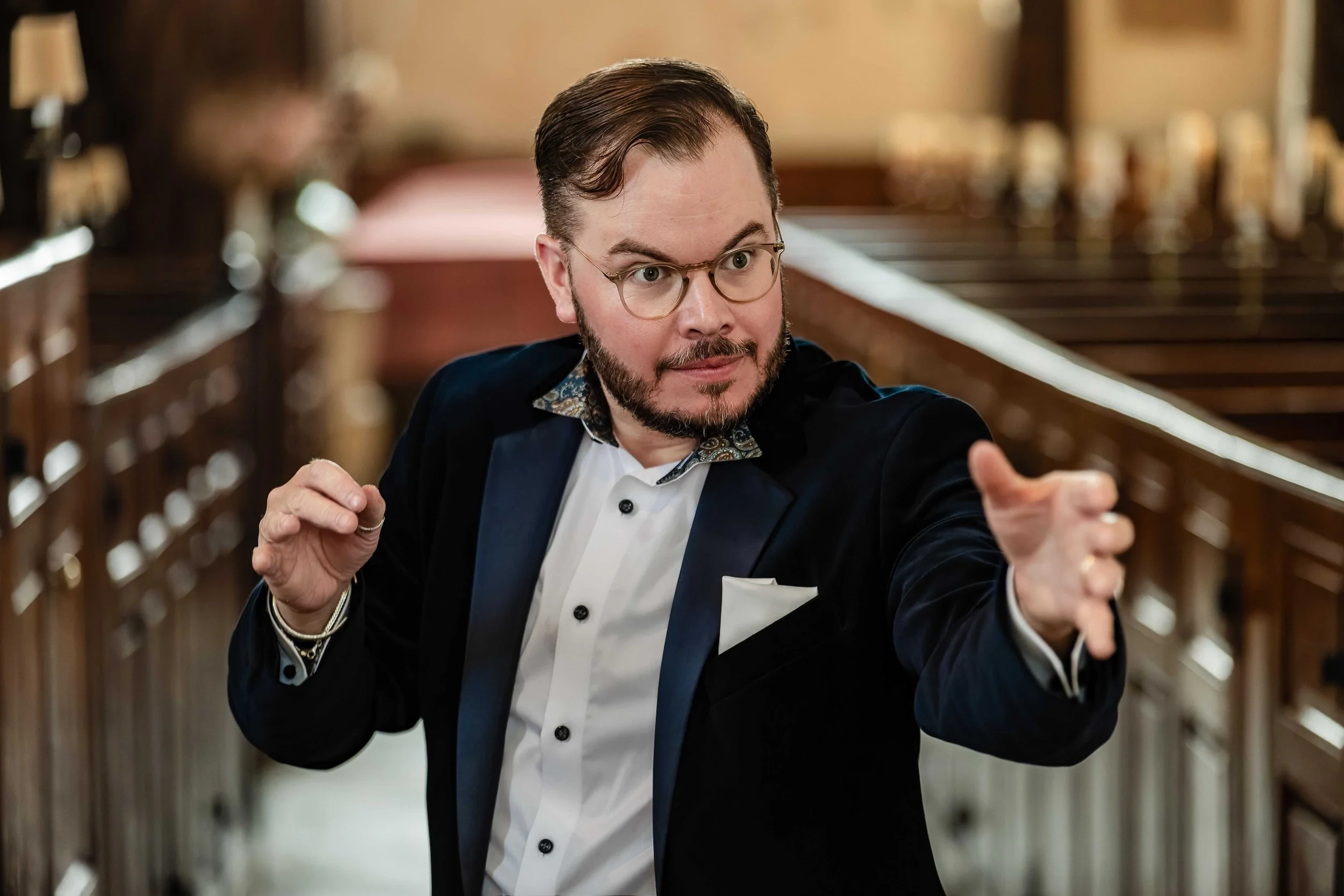 A man in a tuxedo and glasses points with his right hand while looking to his left inside a room with wooden railings and warm lighting.